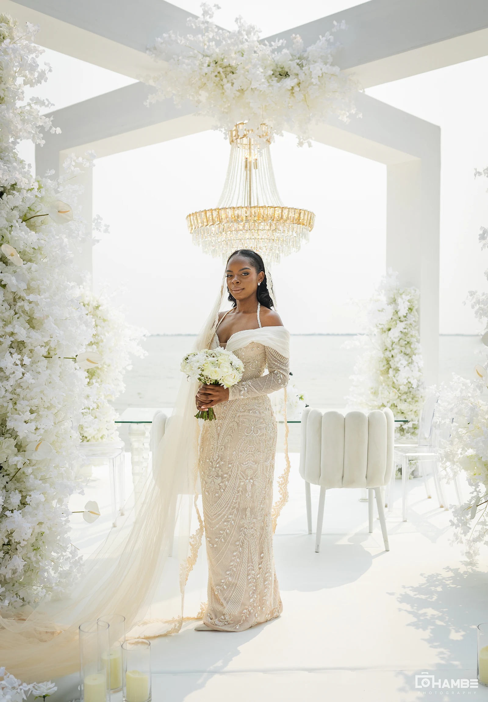 Noiva de vestido bordado e véu comprido sob o arco branco, com ramo branco e o mar ao fundo