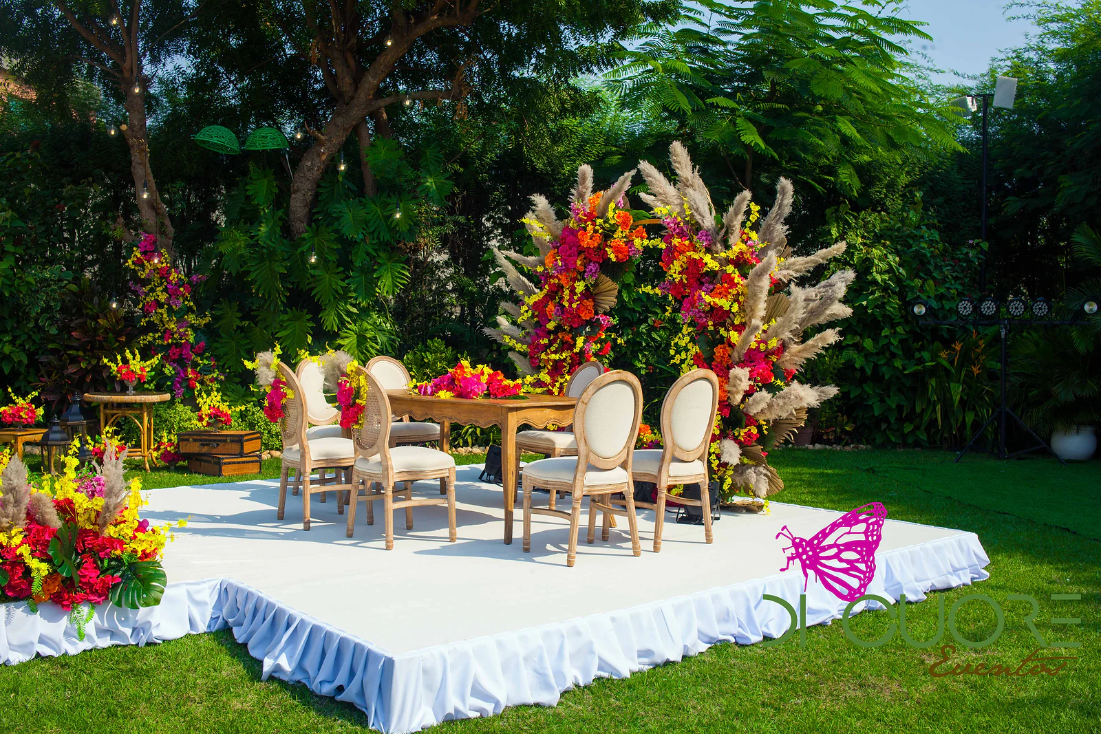 Plataforma branca no relvado, com saia pregueada, mesa de madeira, cadeiras de espaldar redondo e dois aros de flores com erva-das-pampas