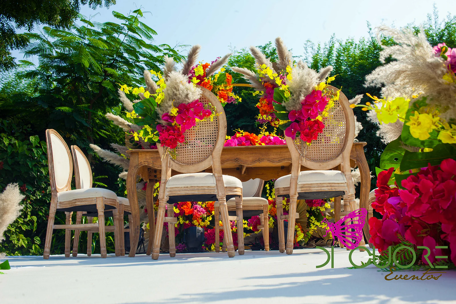 Mesa de madeira entre cadeiras de costas em palhinha enfeitadas com erva-das-pampas e flores fúcsia e amarelas, vistas de baixo