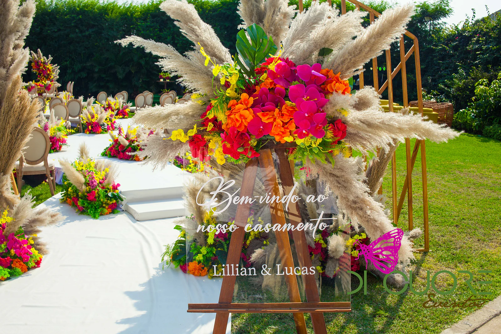Cavalete de madeira com o letreiro «Bem-vindo ao nosso casamento» sob um grande arranjo de flores laranja, fúcsia e amarelas com erva-das-pampas, num jardim com mesas e cadeiras ao fundo