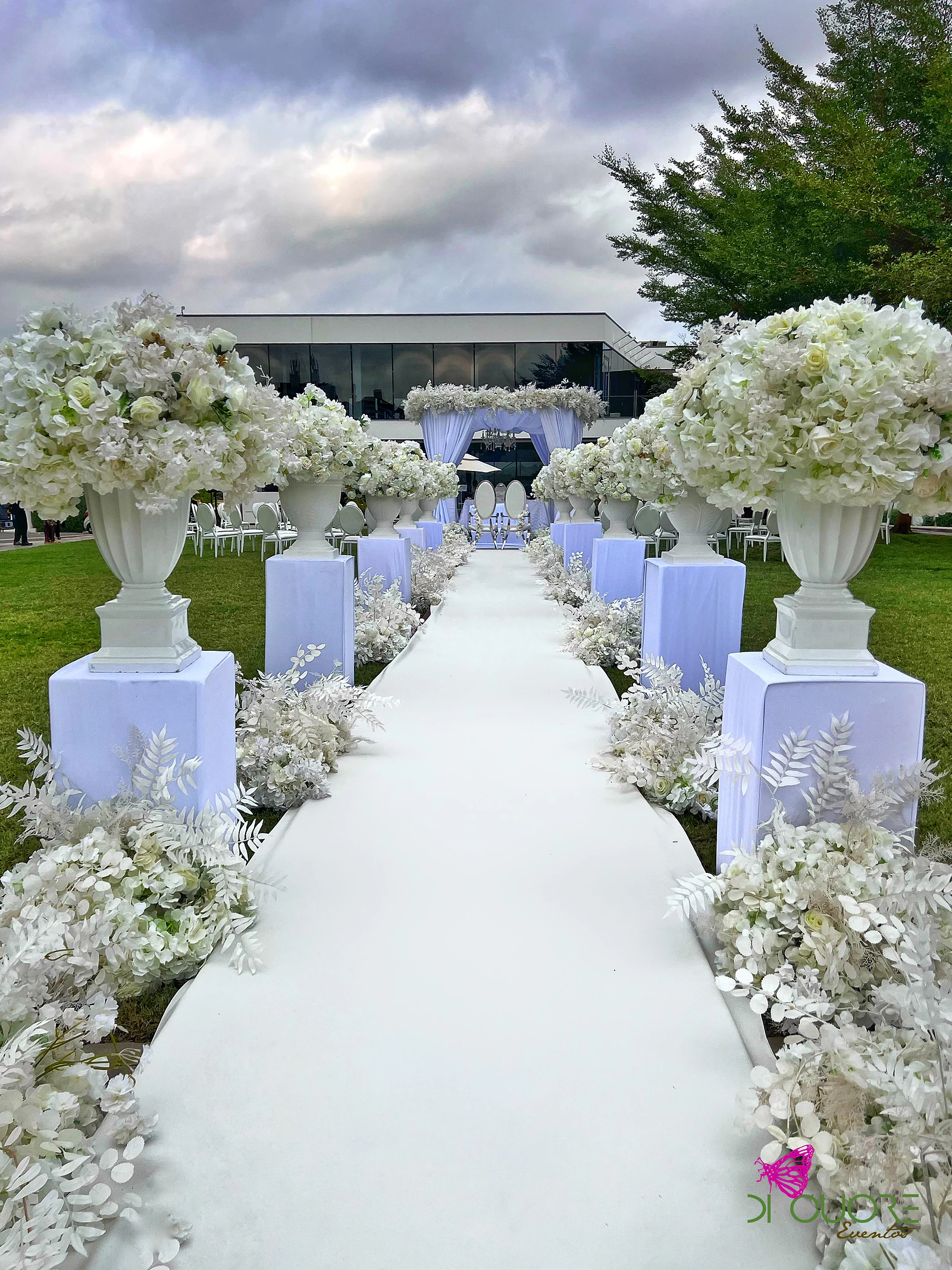 Passadeira branca entre pedestais lilás com urnas de flores brancas e folhagem branca, a conduzir ao pavilhão ao fundo