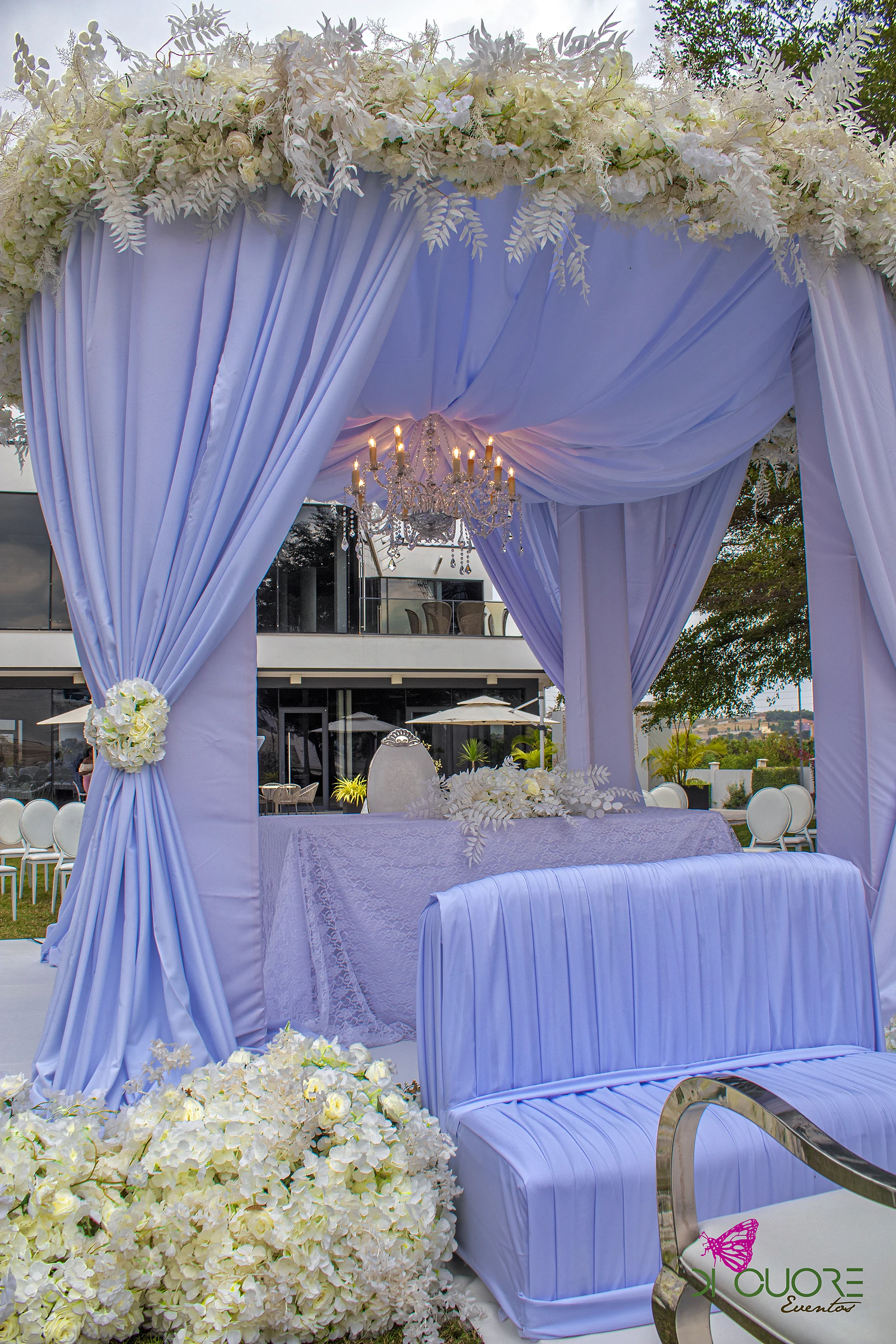Pavilhão de cortinados lilás coroado de flores e folhagem brancas, com um lustre de velas, uma mesa de renda e um sofá lilás em primeiro plano