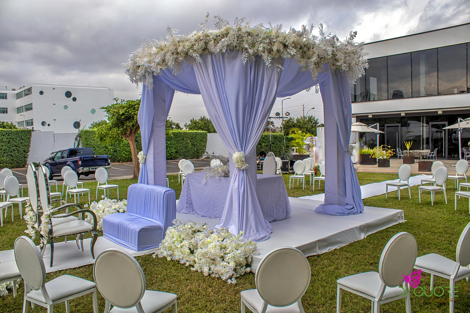 Pavilhão lilás sobre uma plataforma branca no relvado, com cadeiras brancas e maciços de flores brancas à volta