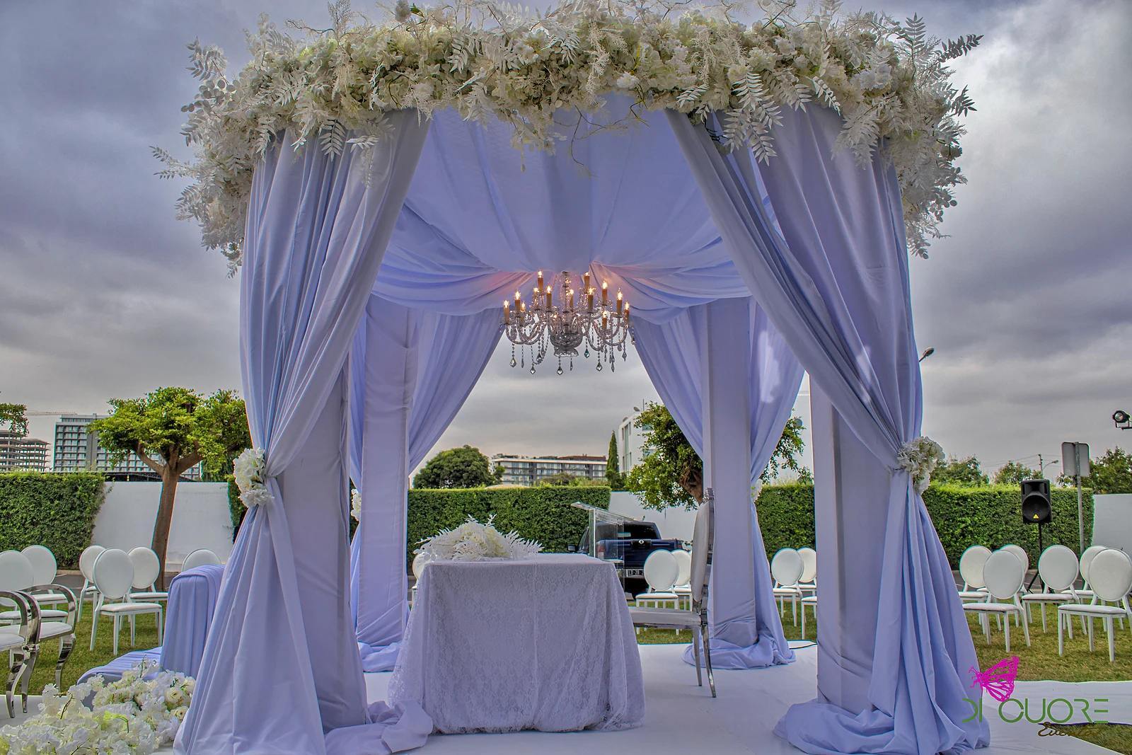 Interior do pavilhão lilás, com cortinados presos por flores brancas, um lustre de velas e uma mesa coberta de renda