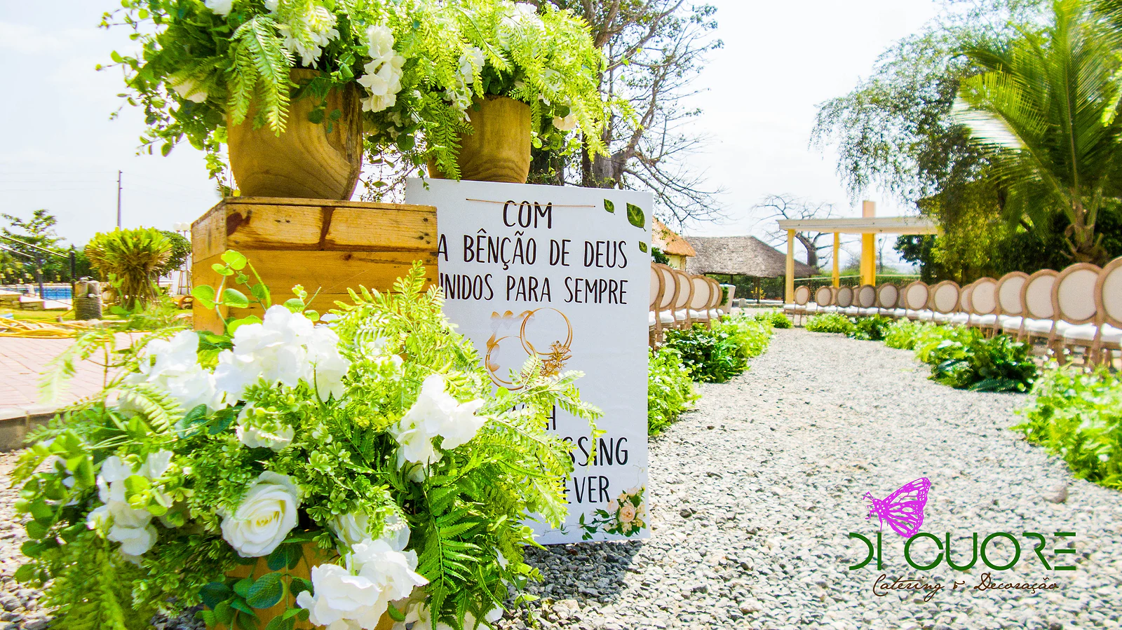 Placa com a frase «Com a bênção de Deus, unidos para sempre» entre rosas brancas e fetos, junto a um caminho de gravilha com cadeiras ao fundo