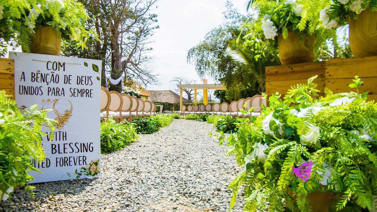 Sign reading “Com a bênção de Deus unidos para sempre” beside pots of white flowers and ferns, at the start of a gravel path lined with wooden chairs