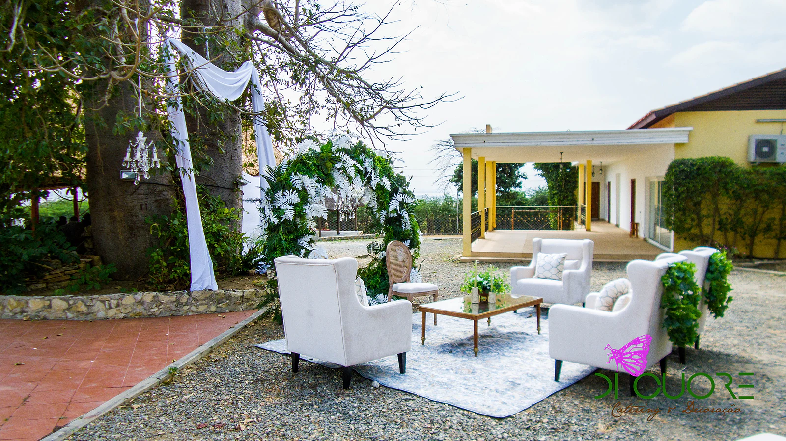 Round arch of foliage and white ferns with a hanging chandelier, a wooden chair and a glass coffee table with white flowers on a blue rug