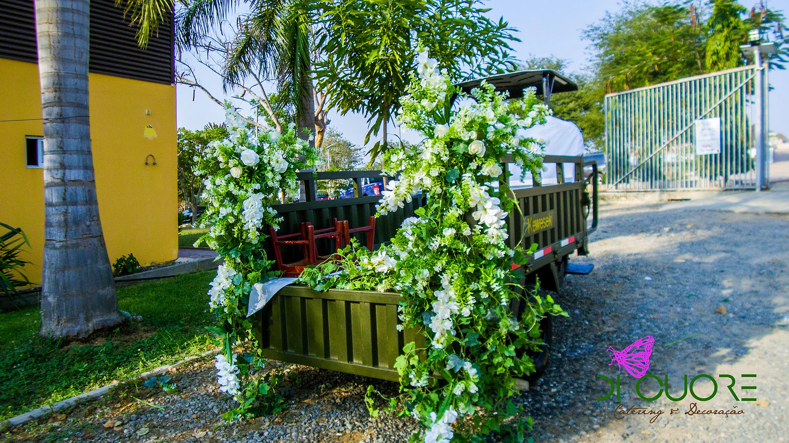 Olive-green cargo vehicle covered in white flowers and foliage, beside a yellow wall and palm trees