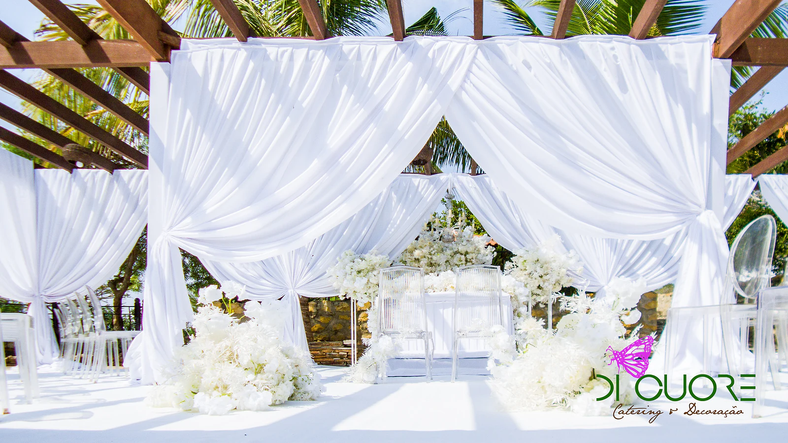 Wooden pergola with tied-back white drapes and, beyond, the altar among white flower arrangements and clear chairs