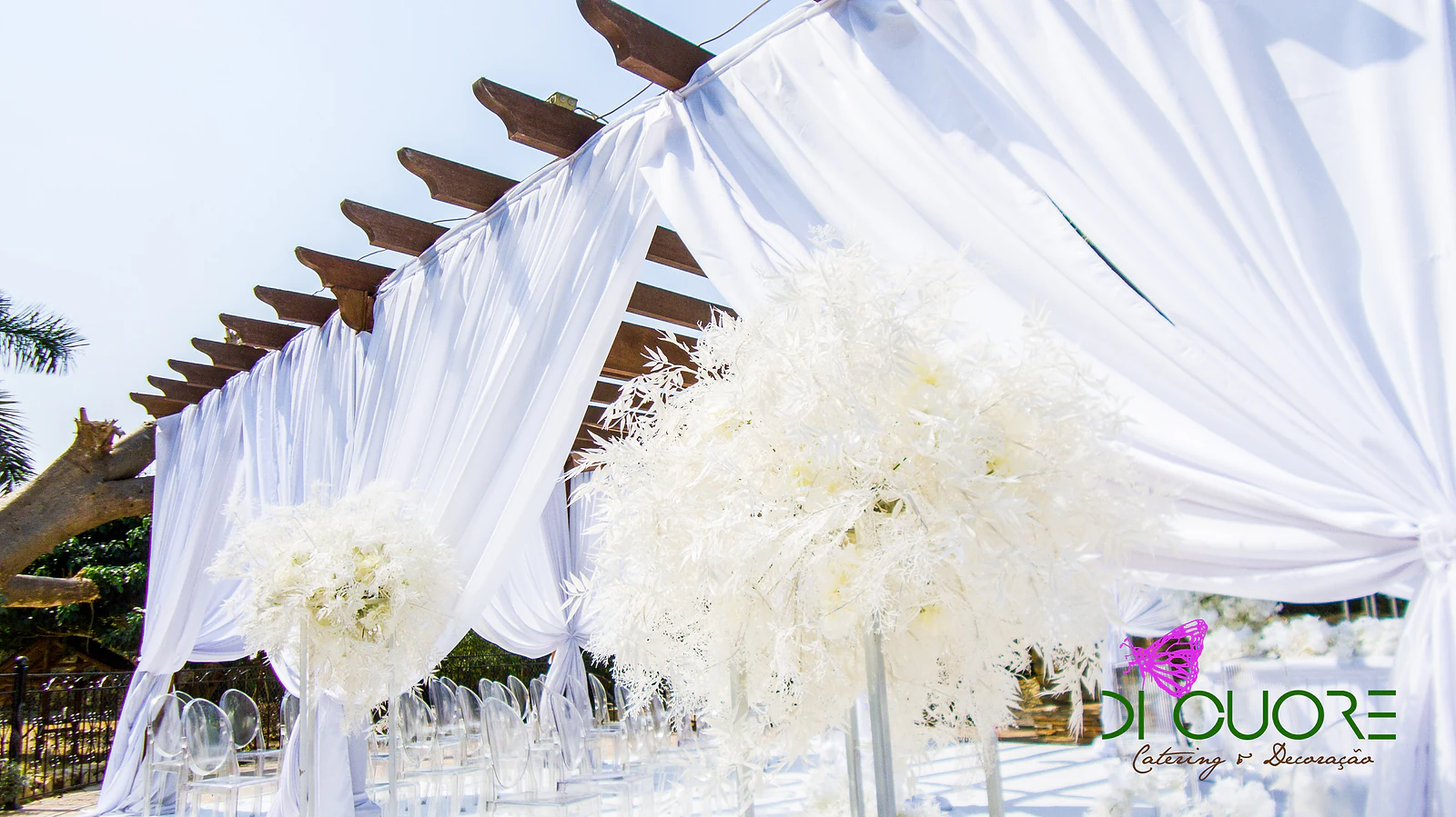 Large balls of white leaves and flowers on clear stands, beside white drapes tied to a wooden pergola