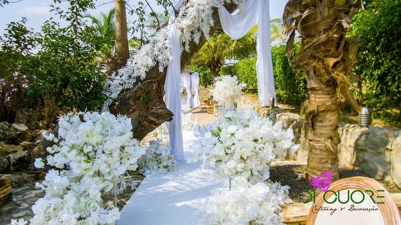 White aisle lined with large arrangements of white flowers, with drapes tied to a trunk and a branch of white leaves