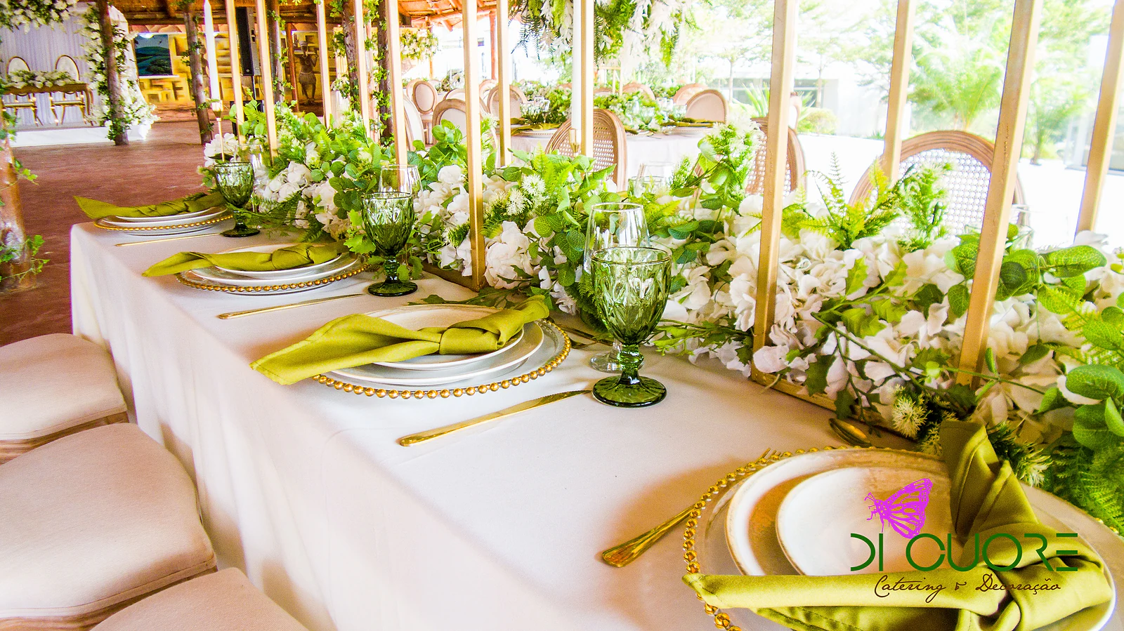 Laid table with gold under-plates, olive-green napkins, green goblets and a garland of white flowers and foliage between golden uprights