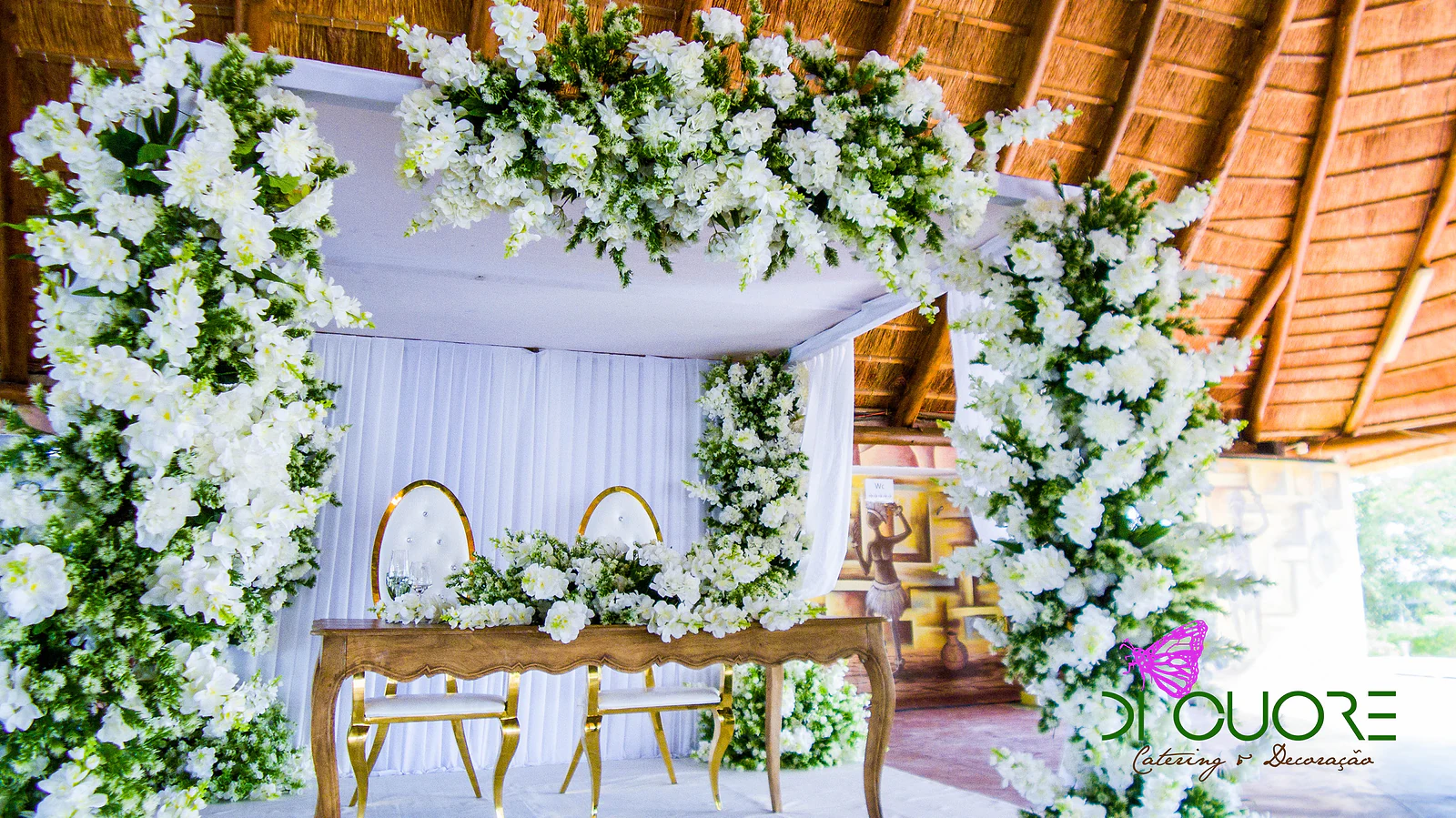 Wooden table with two gold-backed chairs, framed by columns and an arch of white flowers and foliage, beneath the thatched roof