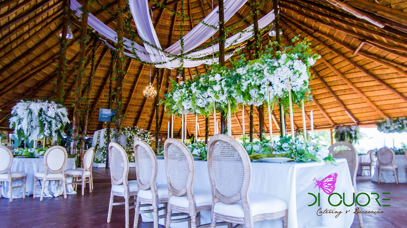 Thatched hall with a long table and a cascade of white flowers hung above tall stands, with drapes on the ceiling and vines on the posts