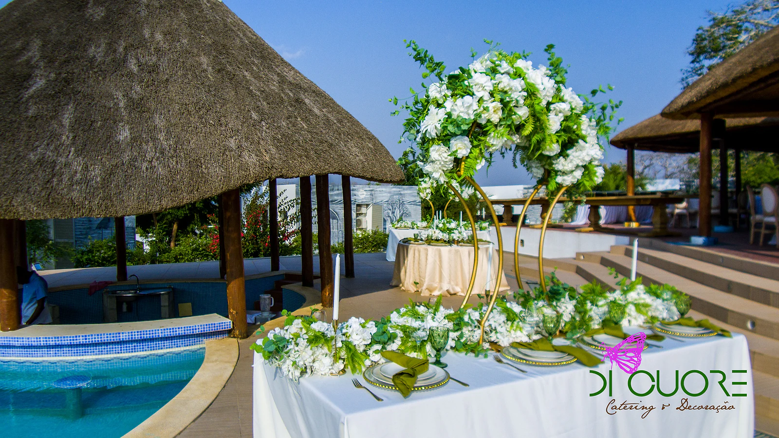 Table with garlands of white flowers and foliage on golden hoops and green napkins, beside the pool and a thatched shelter