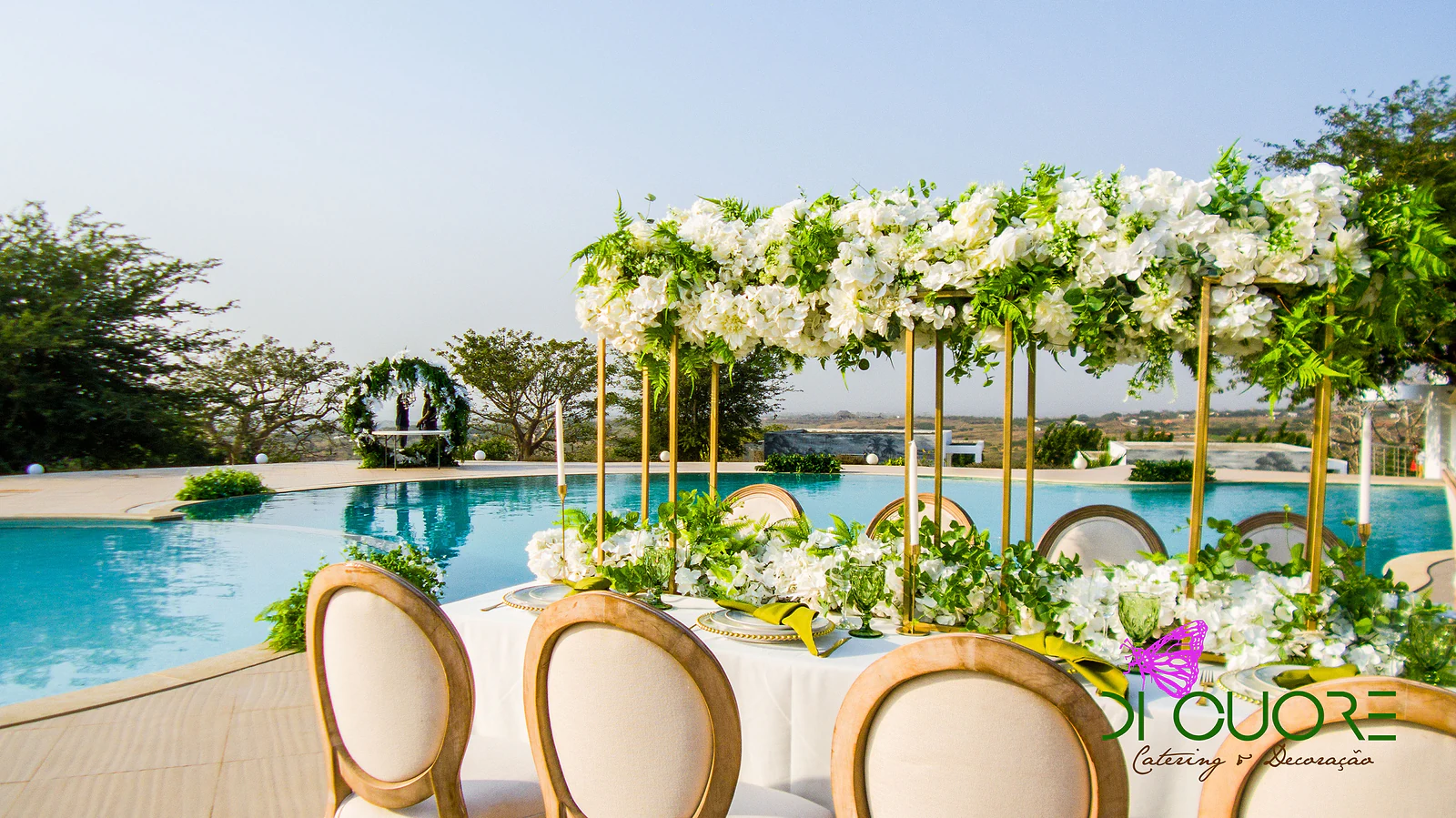 Poolside table with a golden frame covered in white flowers and foliage, wooden chairs and, in the distance, a ring of foliage