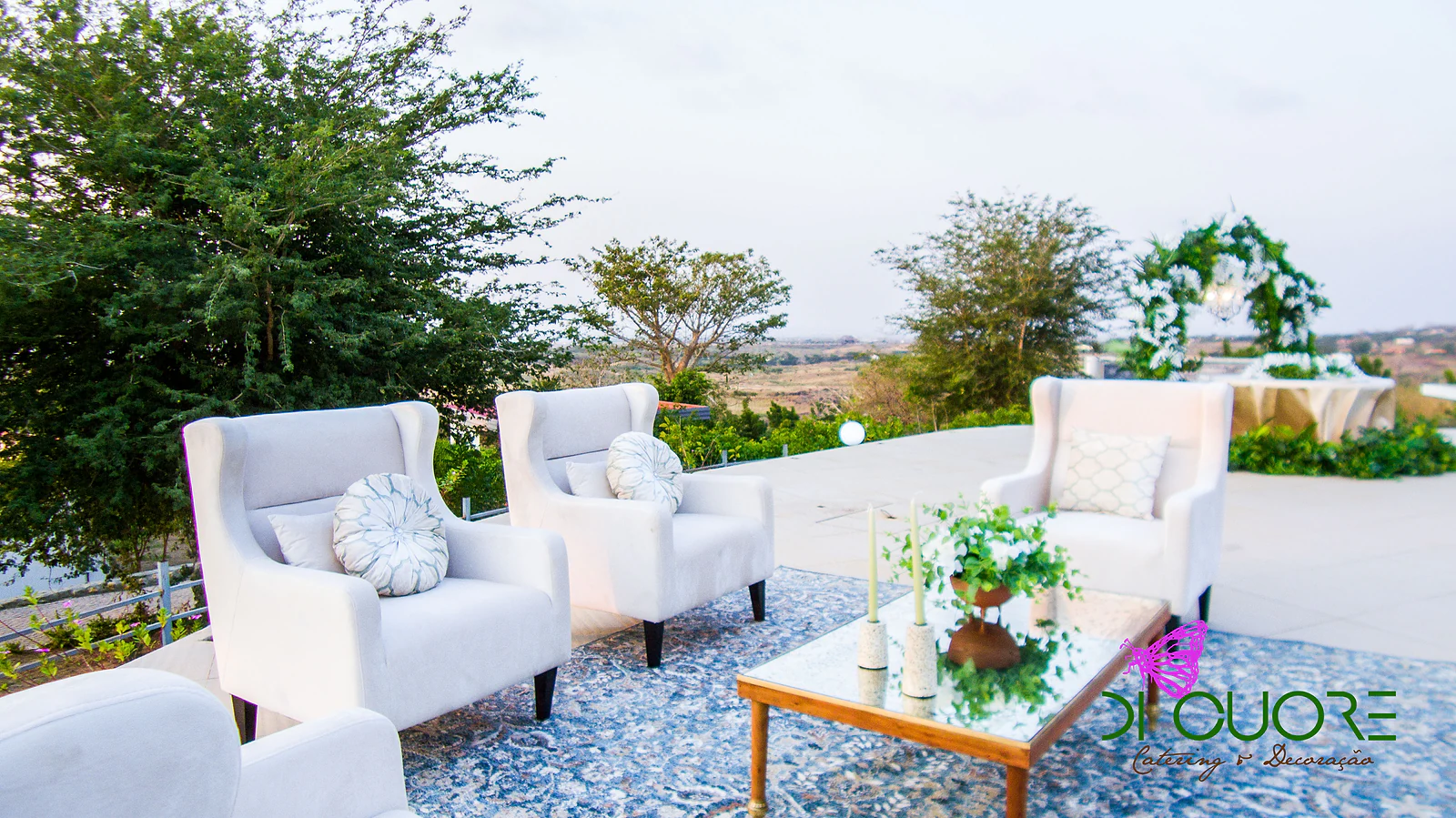 White armchairs with cushions and a glass coffee table with white flowers and candles, on a blue rug, on the terrace