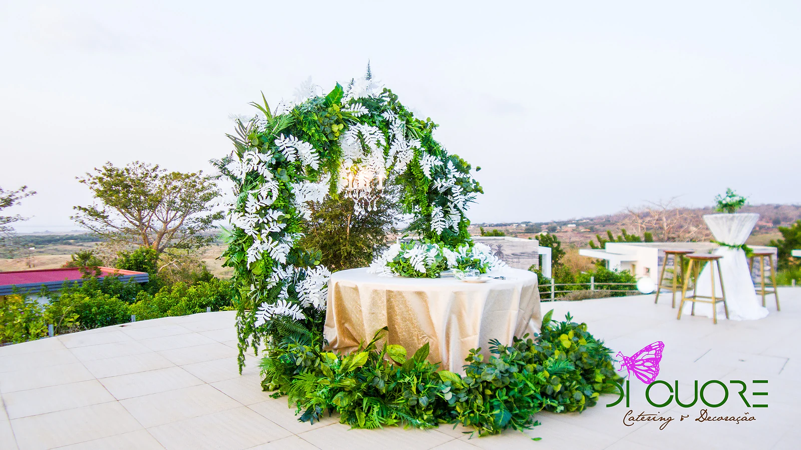 Round arch of foliage and white ferns over a round table in a gold cloth, on a terrace overlooking the landscape