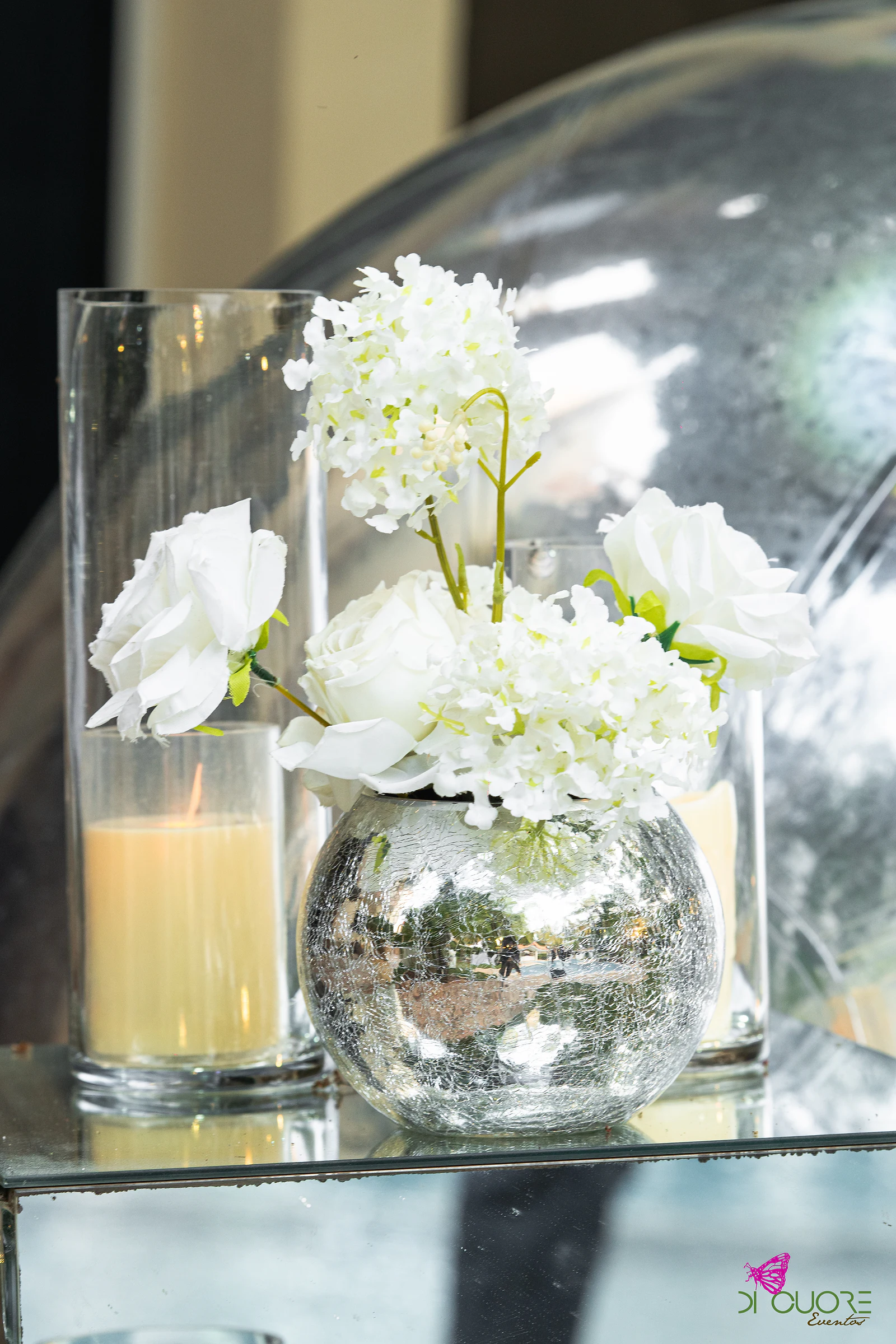 Rosas brancas e flores miúdas num vaso de vidro espelhado, ao lado de uma vela em copo de vidro, sobre um cubo espelhado