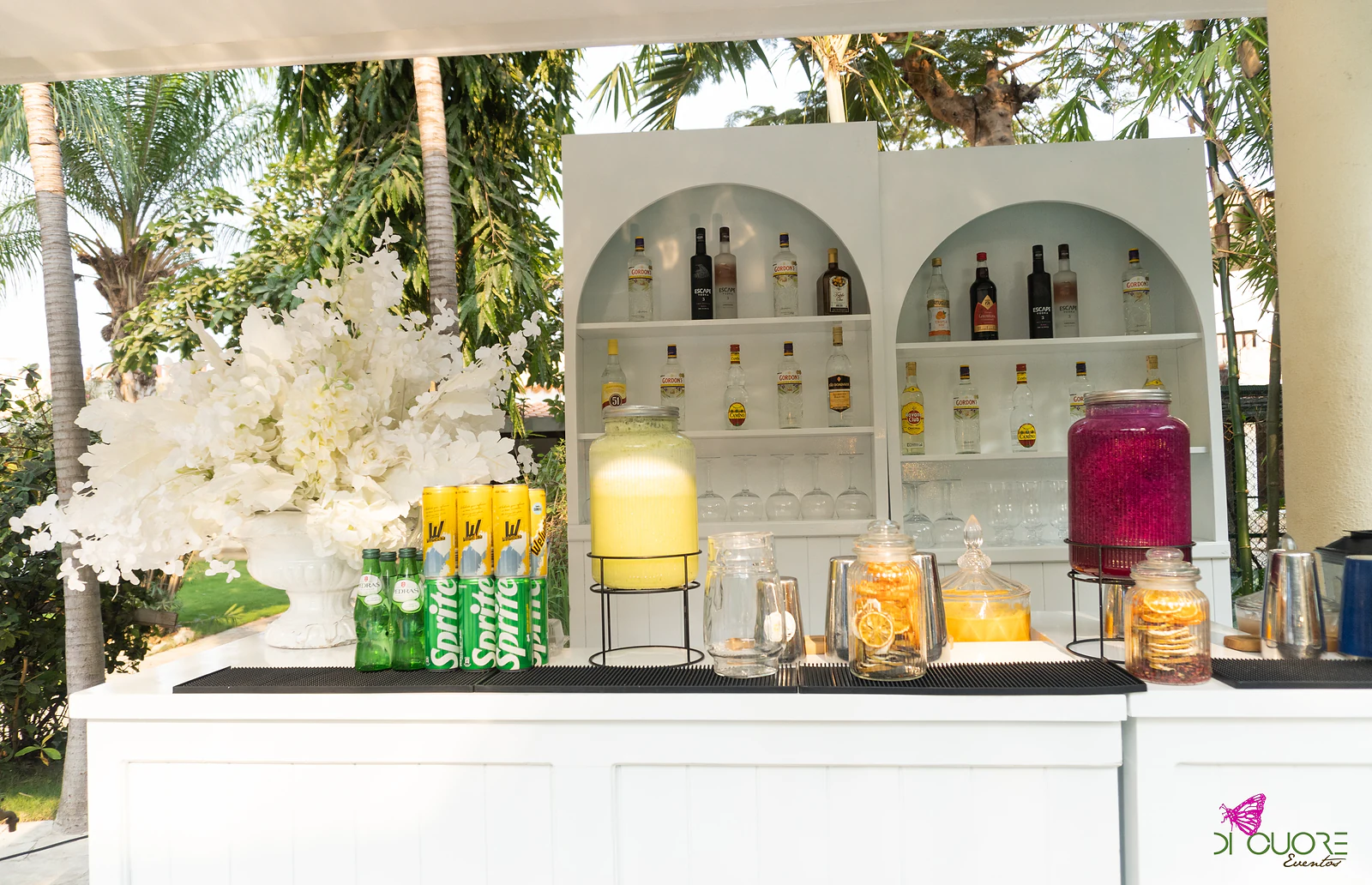 Bar branco com prateleiras em arco e garrafas, jarros de sumo e fruta, e um grande arranjo de flores brancas