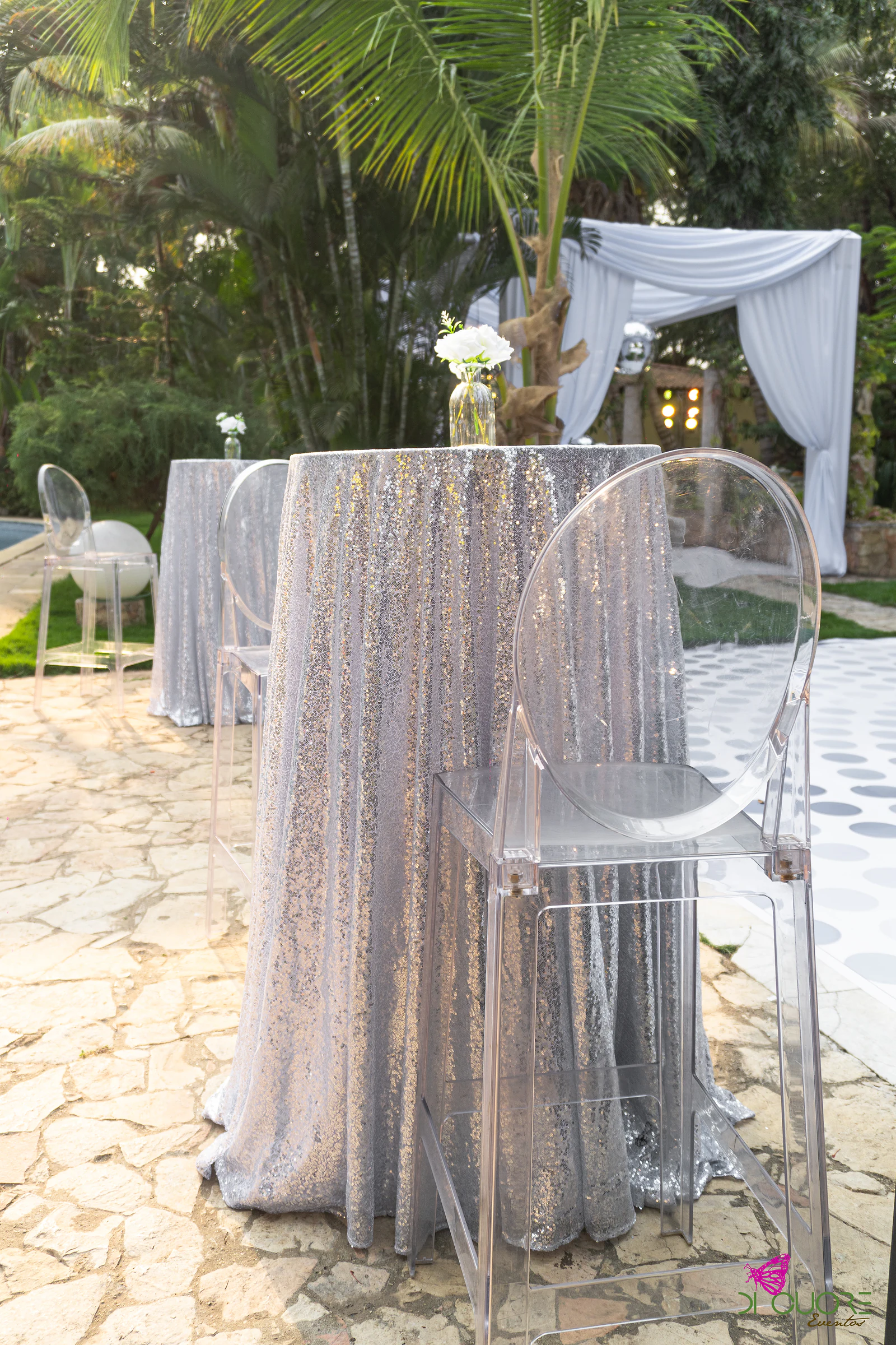 Mesa alta forrada de lantejoulas prateadas, com um pequeno vaso de rosa branca e uma cadeira transparente, num pátio de pedra