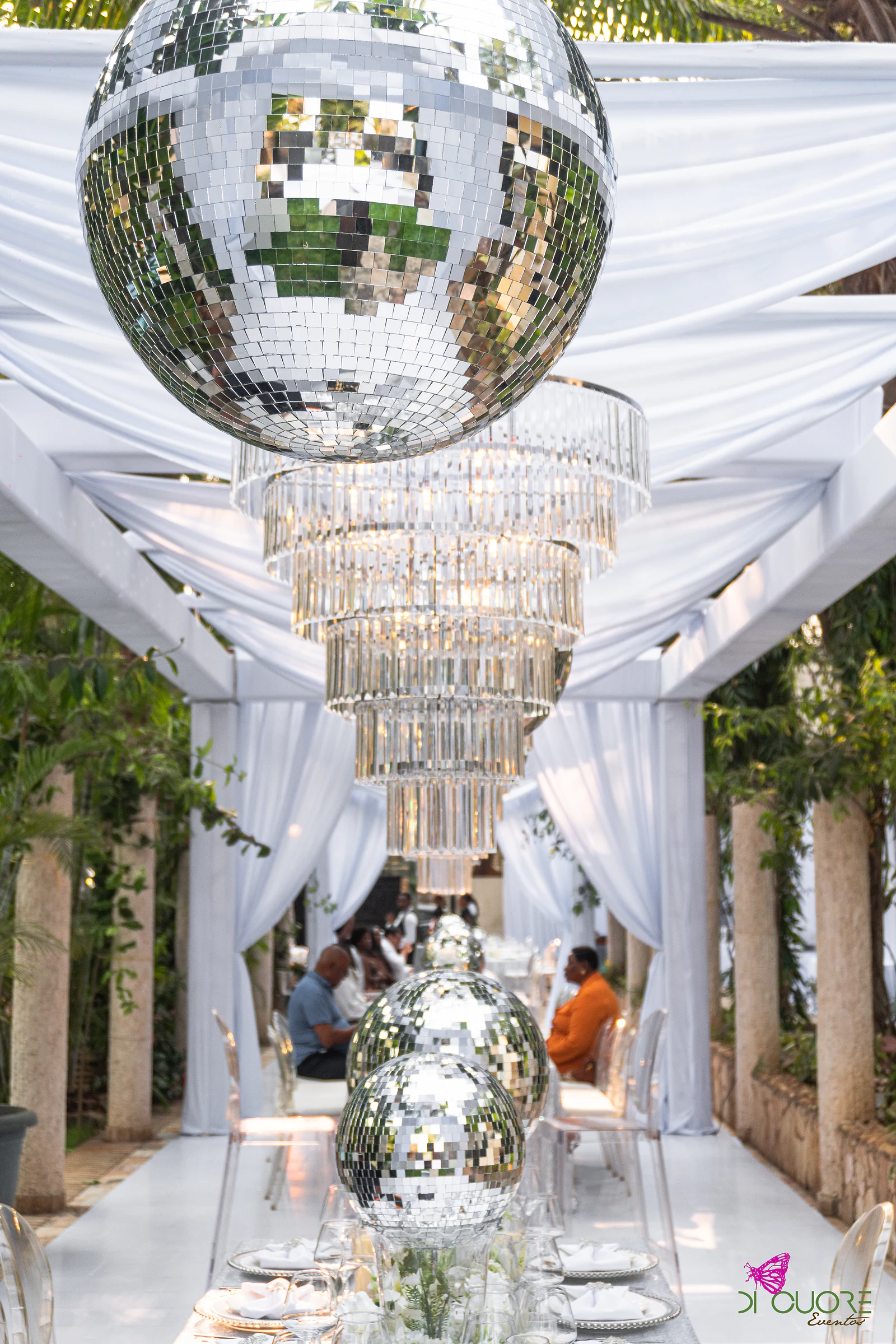 Vista ao longo da pérgola, com uma bola de espelho e um lustre de cristal suspensos sobre a mesa comprida, entre cortinados brancos e colunas