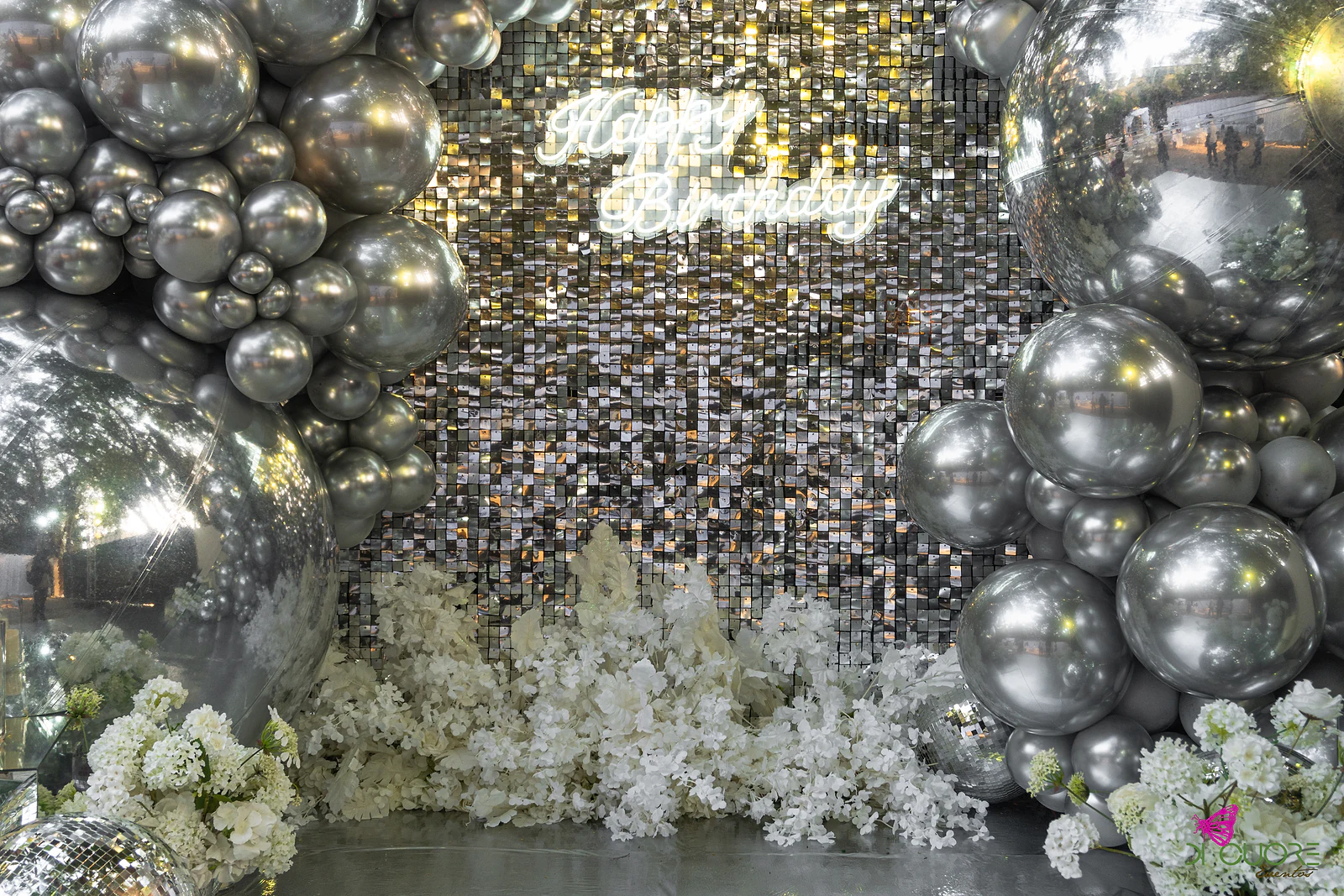 Painel de lantejoulas com o letreiro luminoso Happy Birthday, balões cromados de cada lado e flores brancas junto ao chão