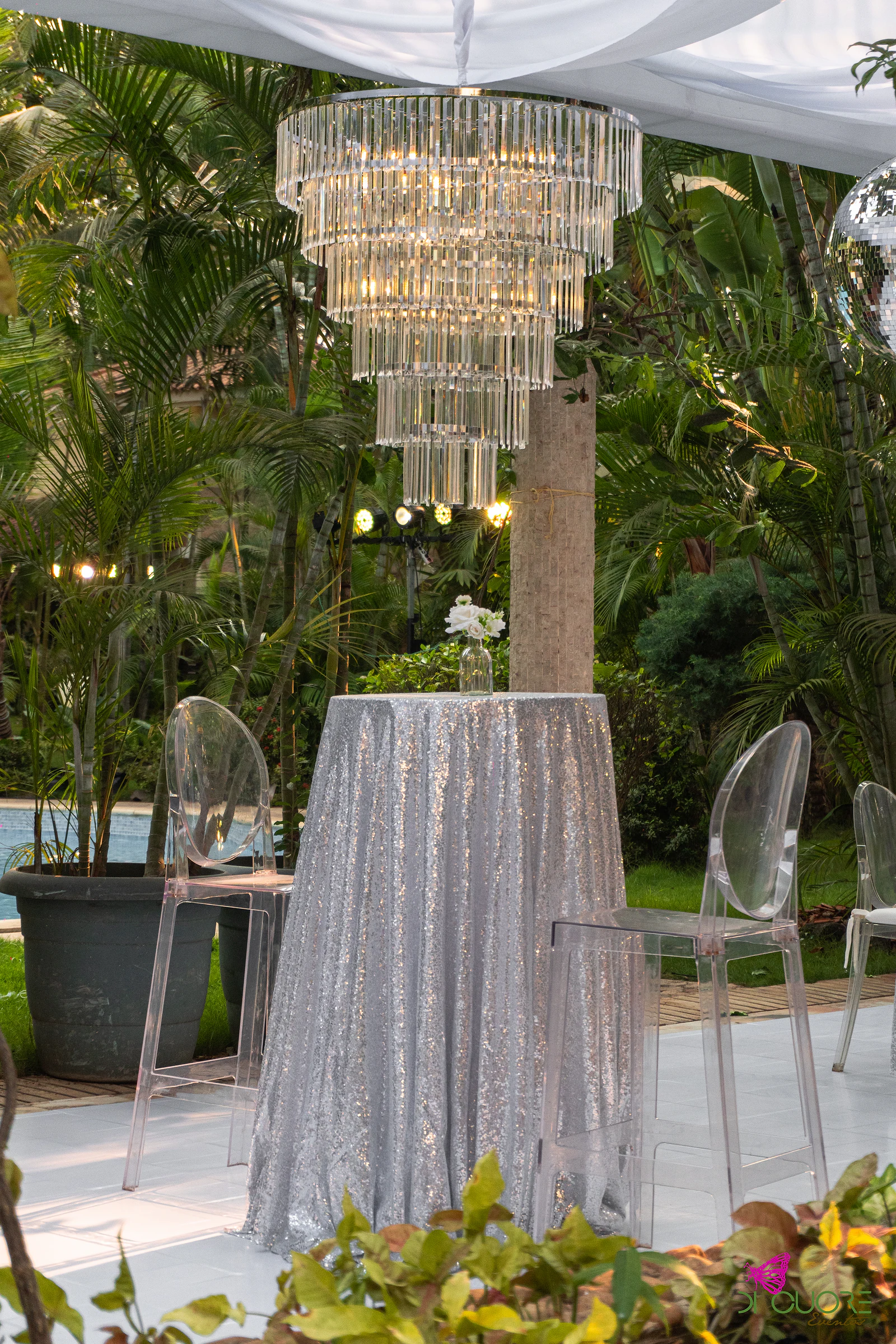 Mesa alta de lantejoulas prateadas com um pequeno ramo de flores brancas e cadeiras transparentes, sob um lustre de cristal e entre palmeiras
