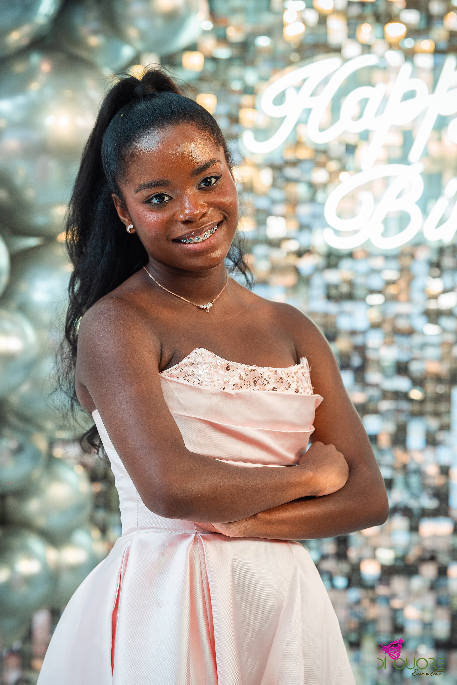 Jovem de vestido rosa-claro sorri diante do painel de lantejoulas com o letreiro luminoso Happy Birthday