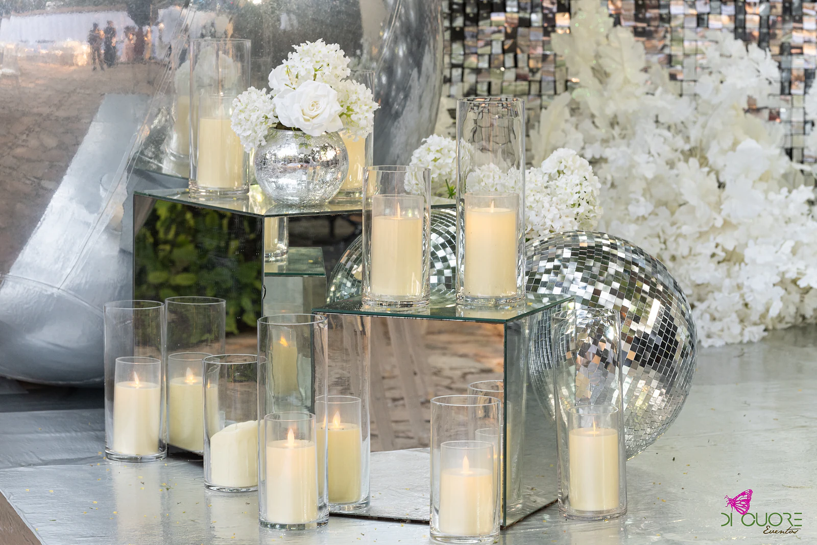 Velas em copos de vidro sobre cubos espelhados, com flores brancas e uma bola de espelho