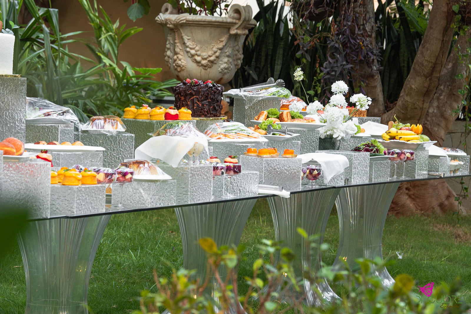 Mesa de doces sobre pedestais texturados, com tartes, pastéis de fruta, taças e flores brancas, num canto do jardim