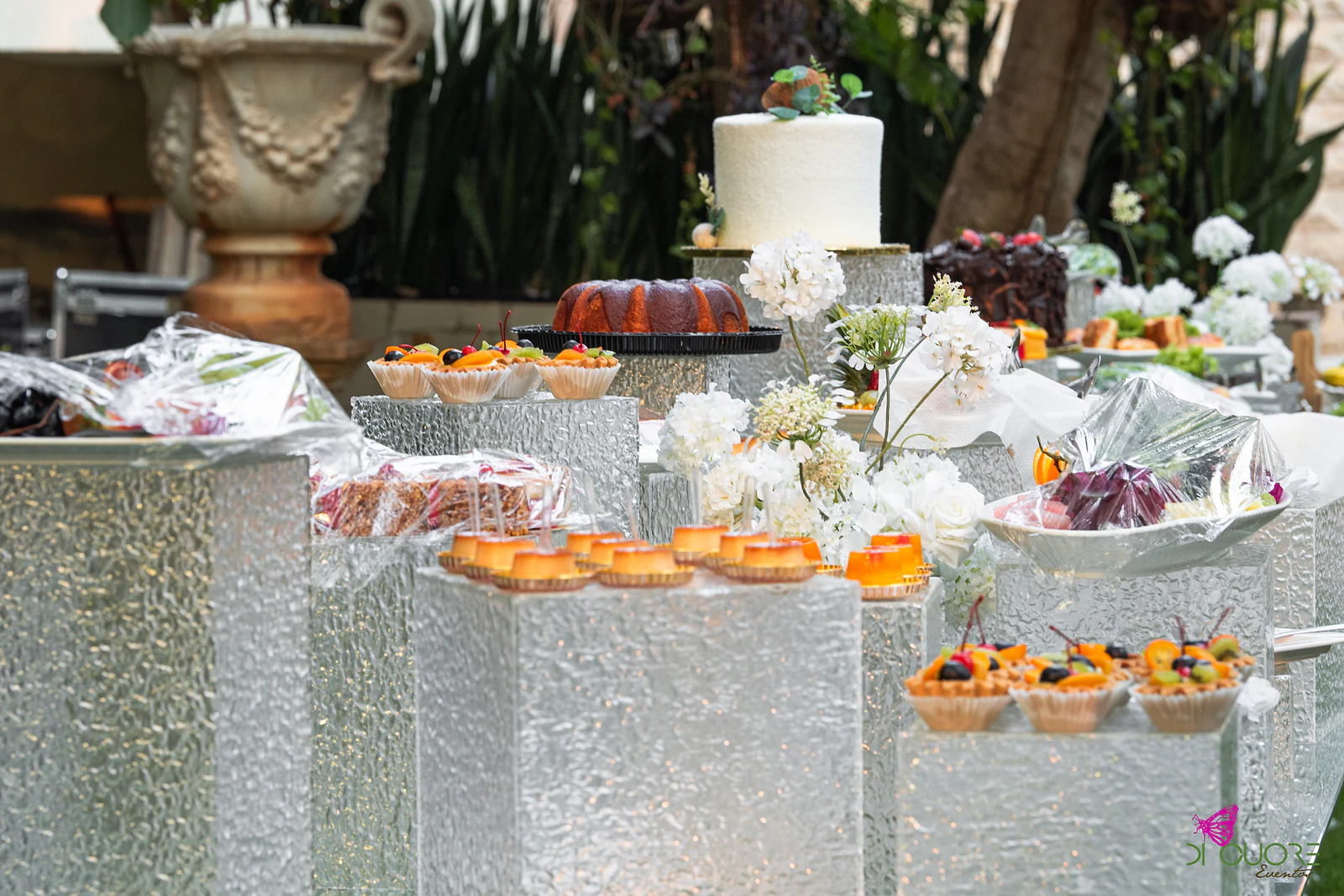 Mesa de doces com pedestais texturados, tartes de fruta e bolo branco ao fundo