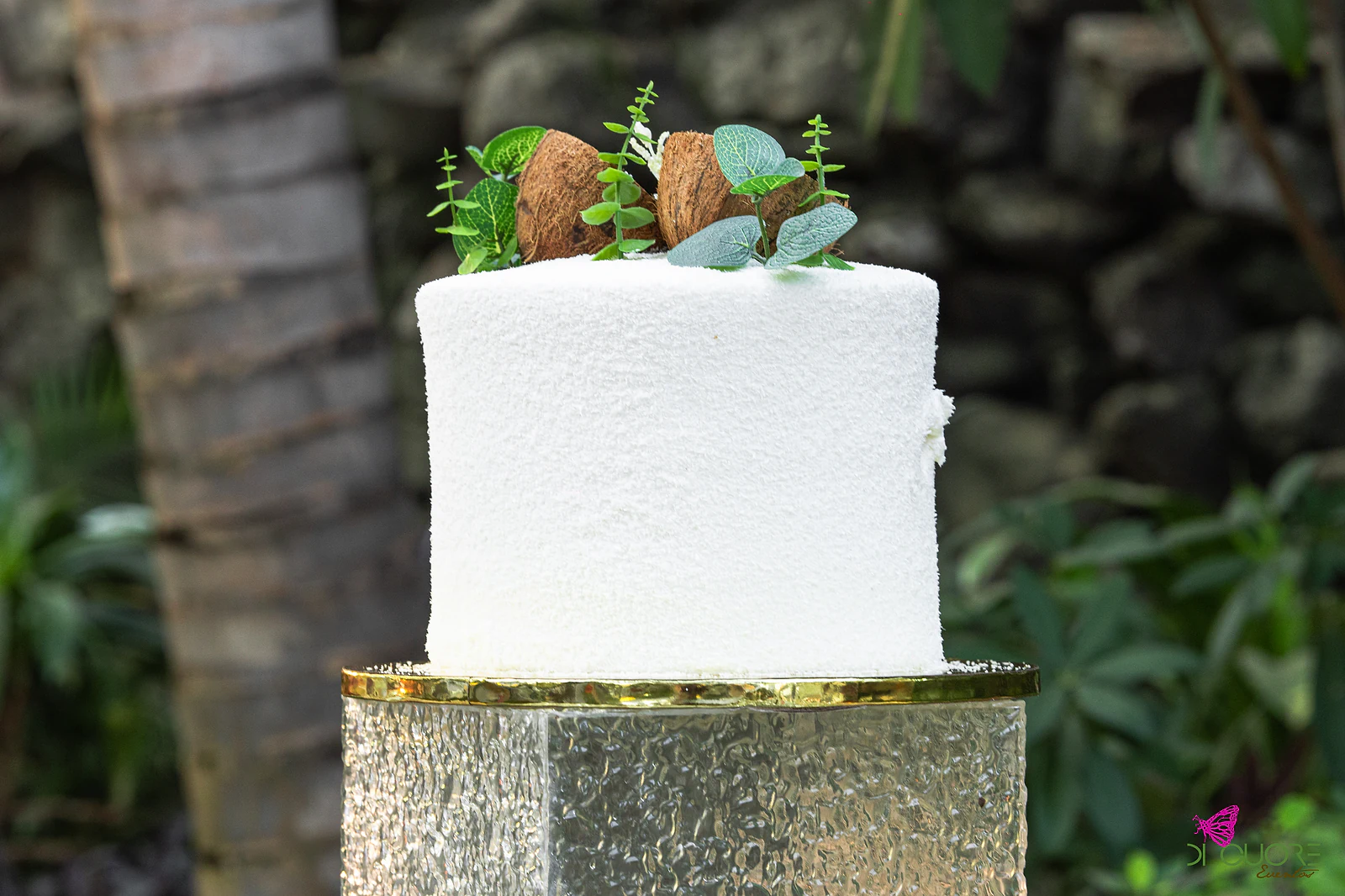Bolo branco texturado, decorado com cocos e ramos de folhagem, sobre um pedestal translúcido