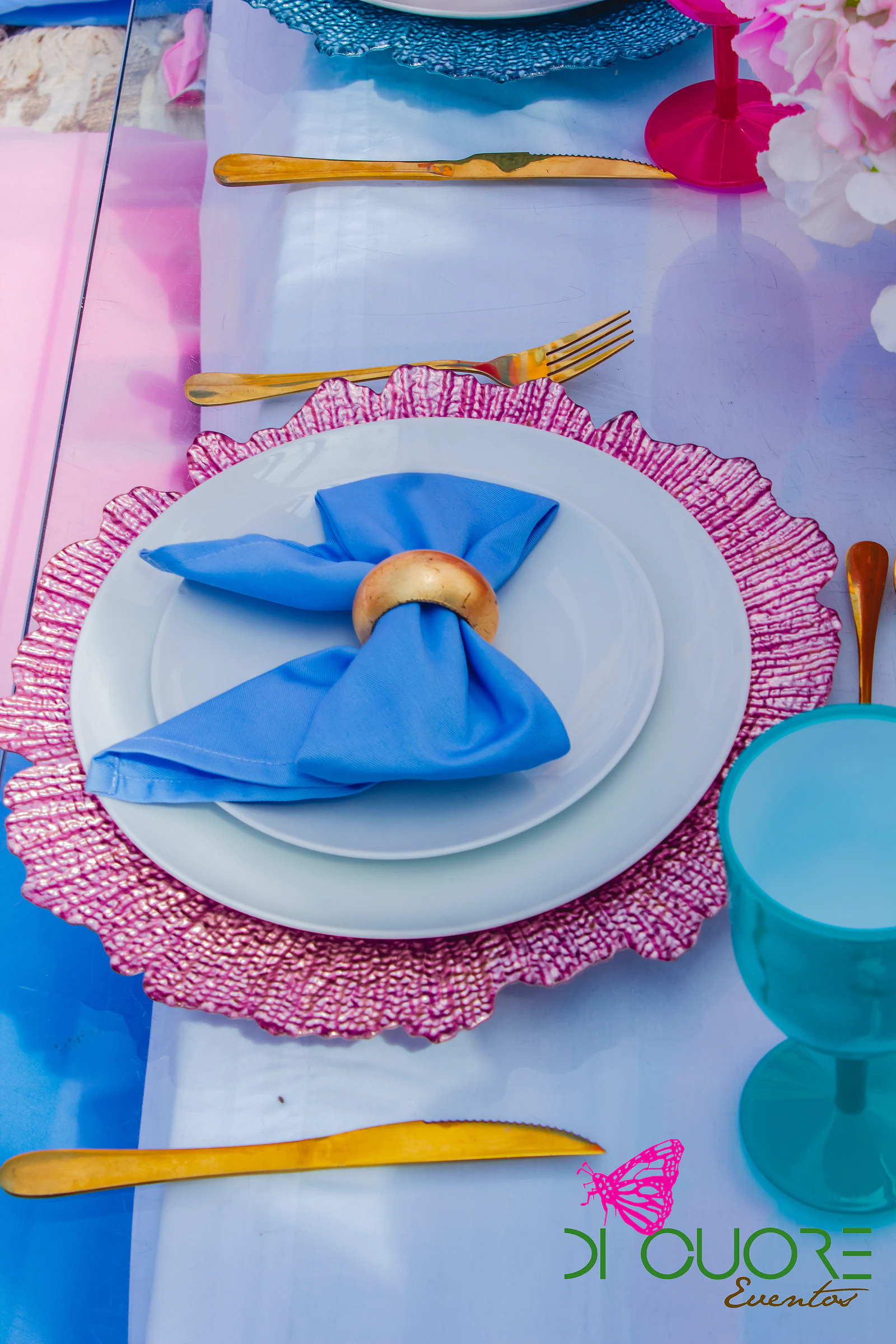 Prato branco sobre sousplat rosa com guardanapo azul em laço e argola dourada, talheres dourados e copo azul-turquesa, vistos de cima