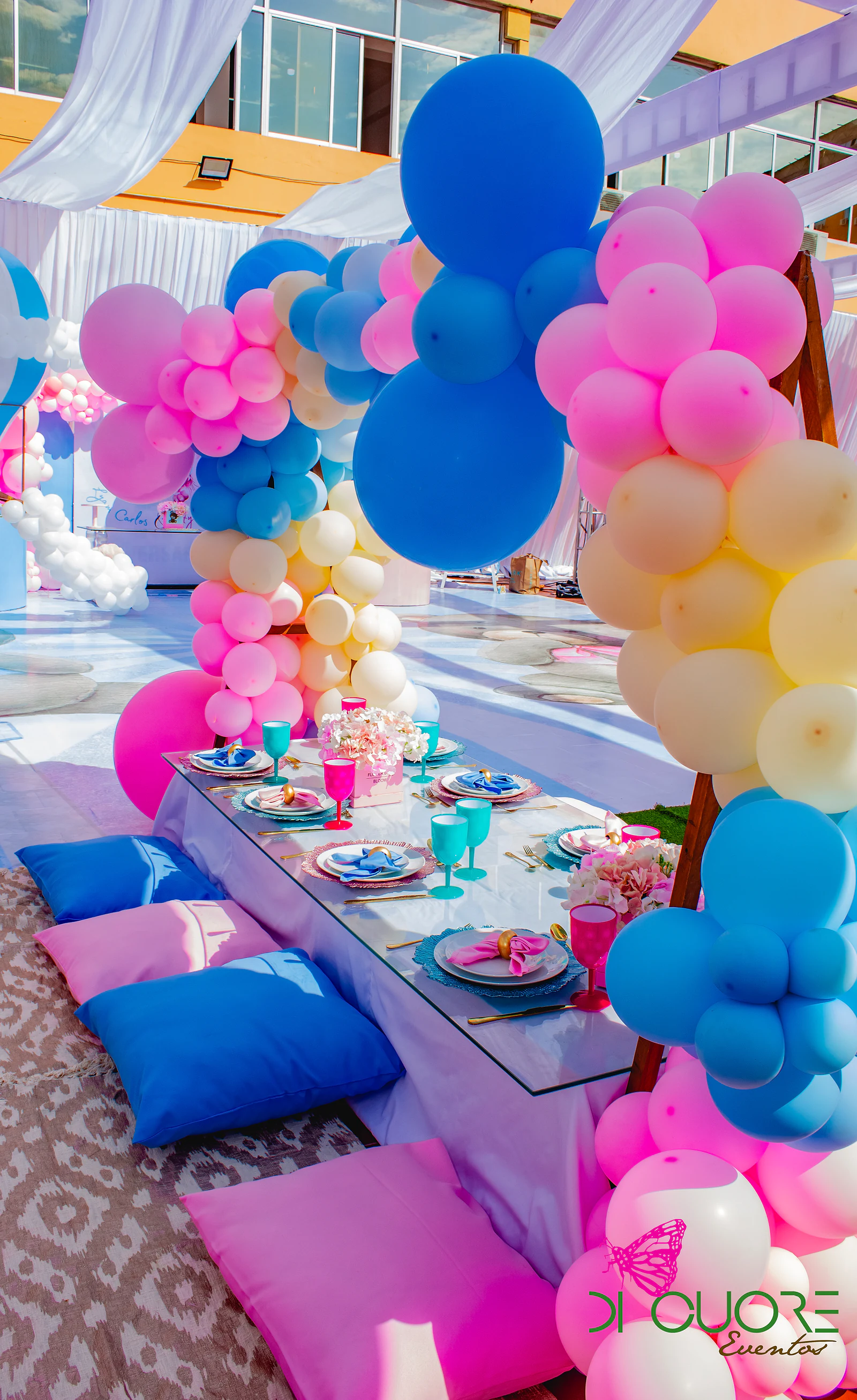 Mesa baixa posta entre almofadas rosa e azuis, com arcos de balões em rosa, azul e creme de cada lado e cortinados brancos por cima