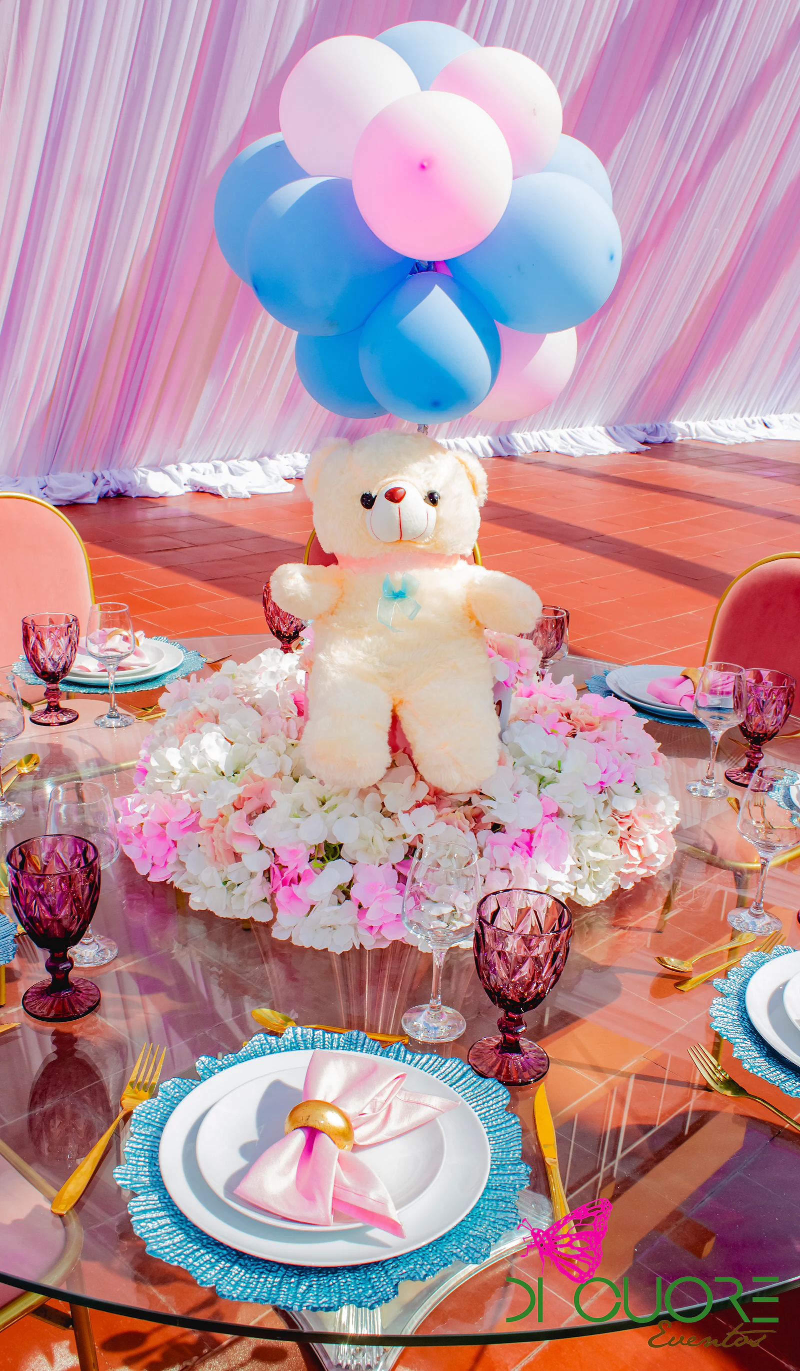 Ursinho de peluche com um cacho de balões rosa e azuis sobre um ninho de flores cor-de-rosa, no centro de uma mesa de vidro posta