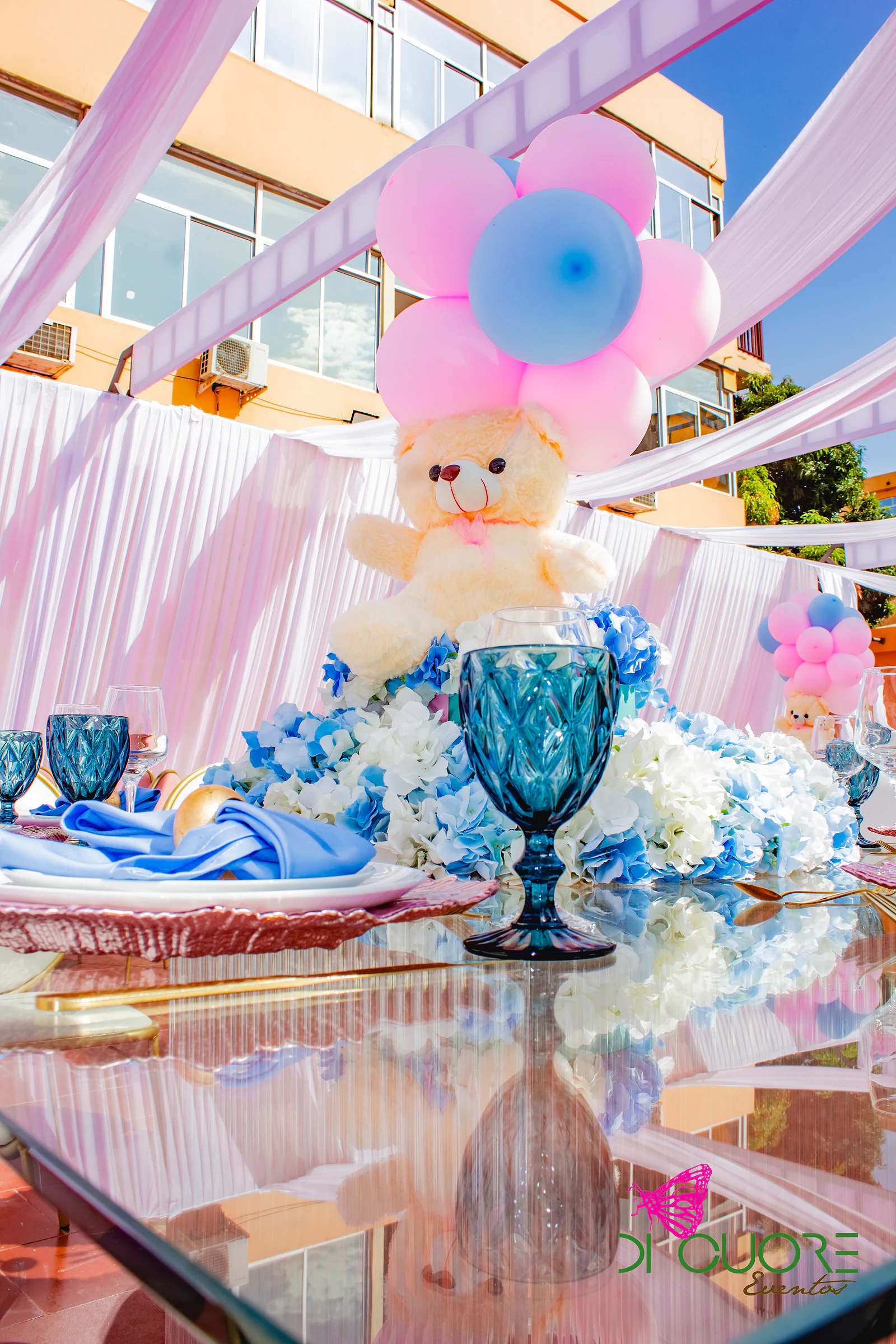 Ursinho de peluche com balões cor-de-rosa e azuis sobre flores azuis e brancas, com um copo azul de vidro lapidado em primeiro plano
