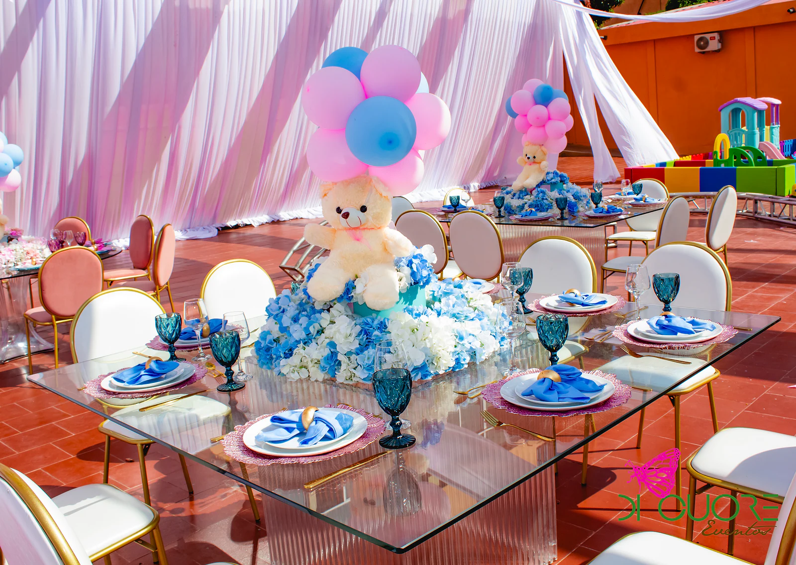Mesa de vidro comprida posta com sousplats rosa e guardanapos azuis, com um ursinho de peluche e balões sobre flores azuis, entre cadeiras brancas e cor-de-rosa