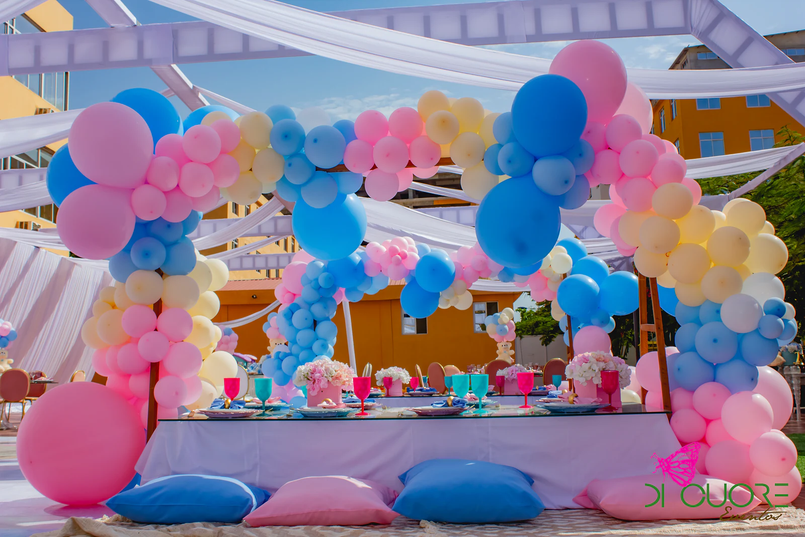 Mesa baixa com almofadas rosa e azuis sob um arco de balões em rosa, azul e creme, com cortinados brancos e céu azul