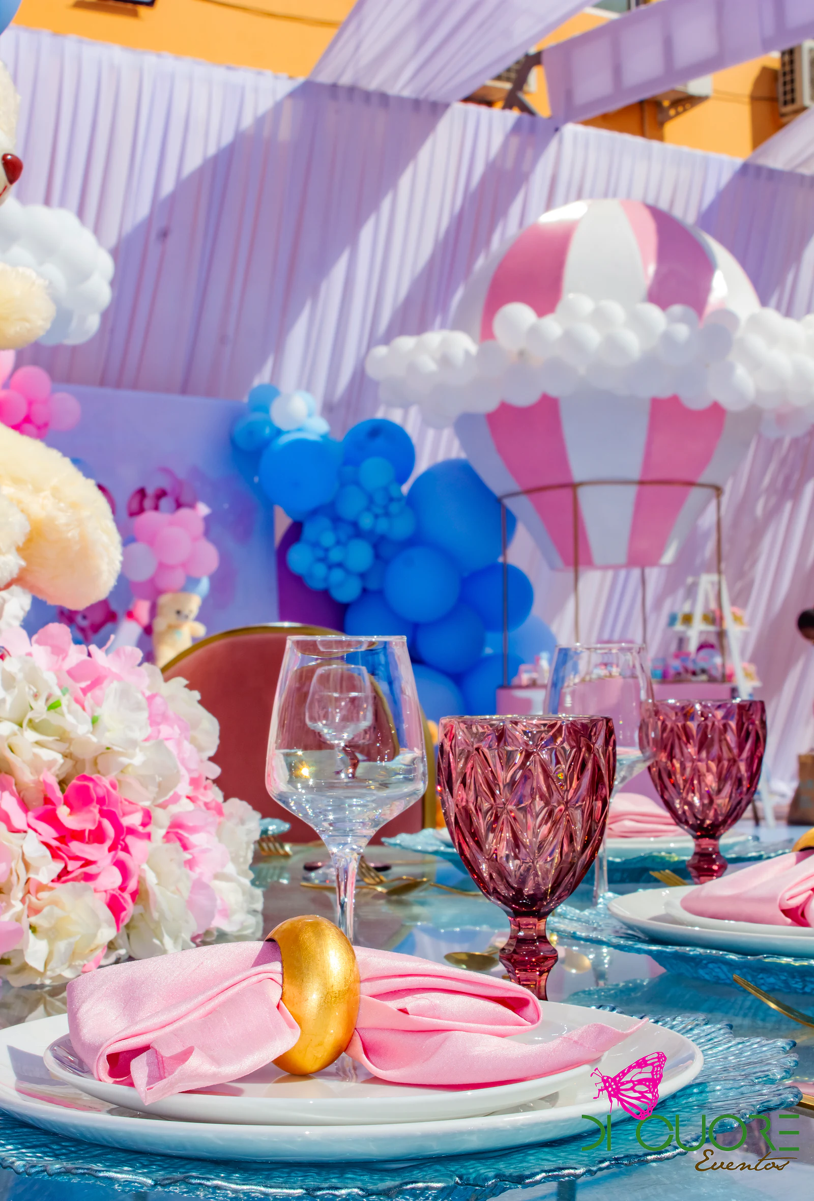 Mesa posta com guardanapo rosa em laço e copos cor-de-rosa, com flores e um balão de ar quente cor-de-rosa e branco ao fundo