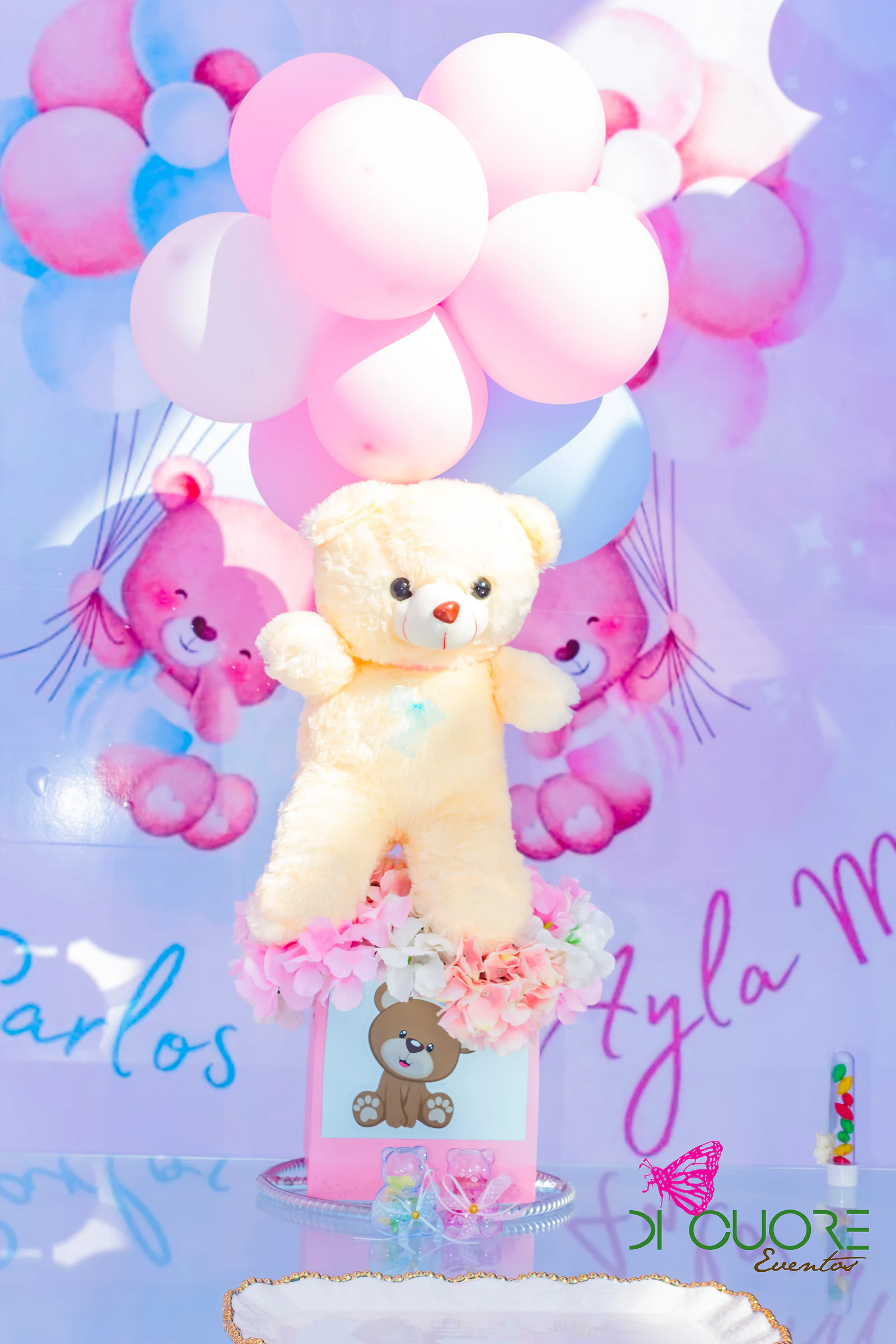 Plush bear tied to a cluster of pink balloons above a box of flowers, before a backdrop of painted bears and handwritten lettering