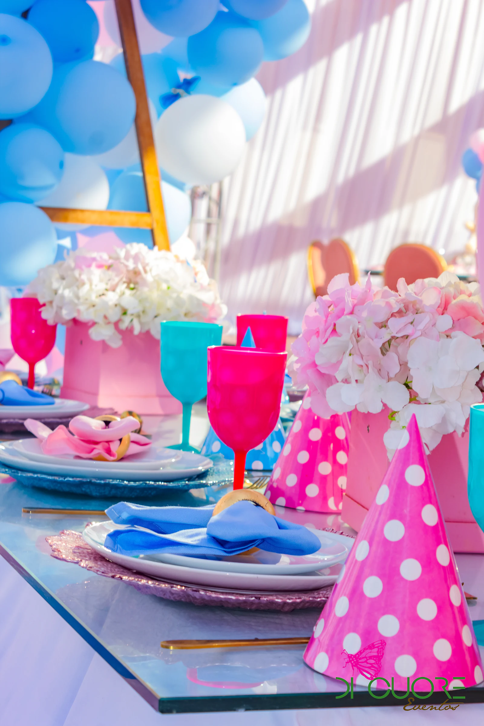 Mesa de vidro com chapéus de festa cor-de-rosa de bolinhas brancas, copos fúcsia e azul-turquesa, flores e um guardanapo azul em laço