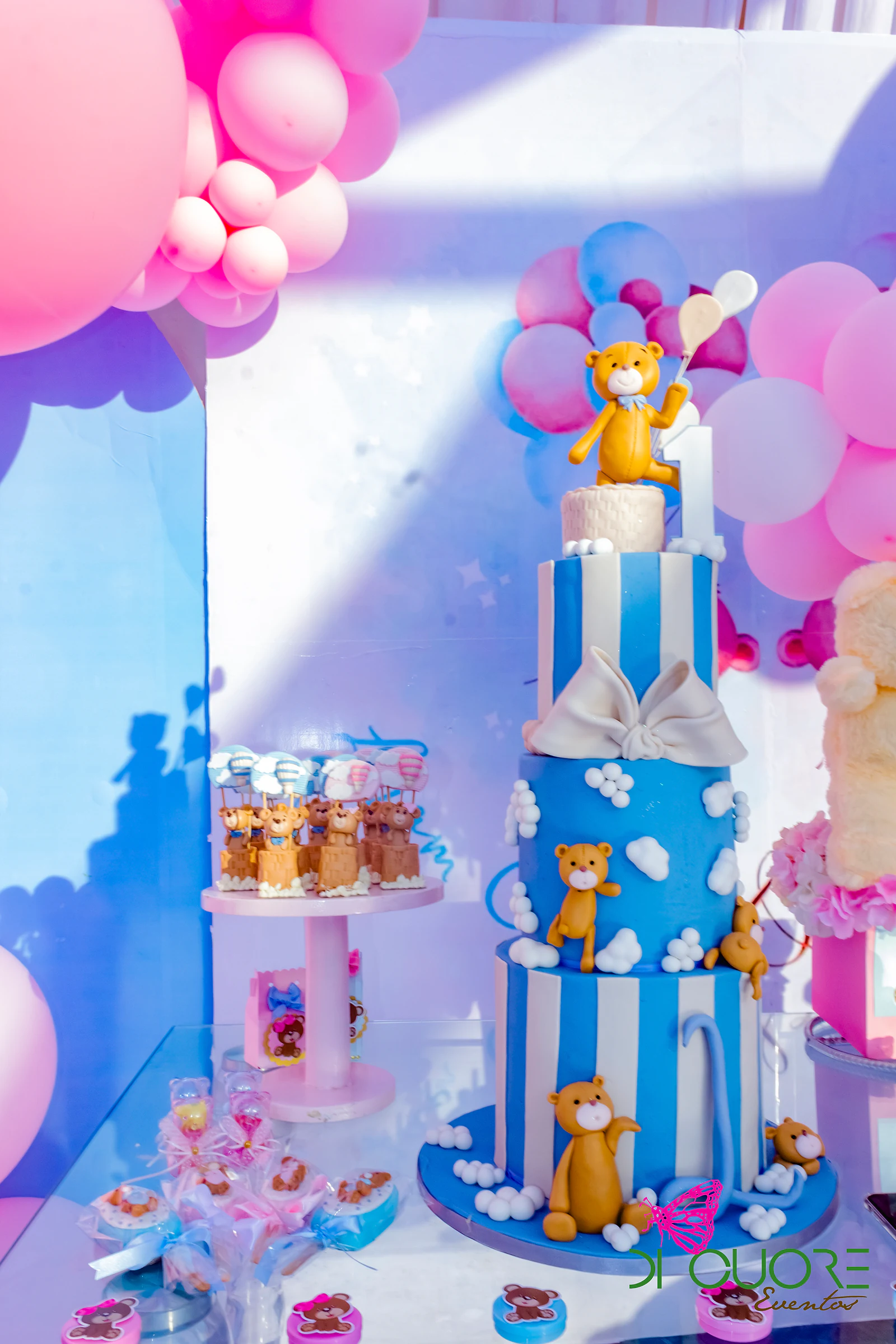Bolo azul de listas brancas com ursinhos de massa e um laço, junto de doces e de um painel de balões