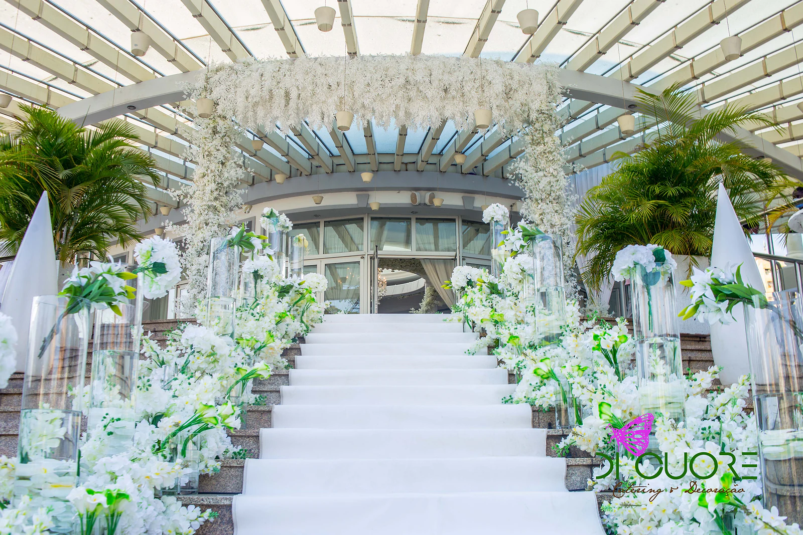 Escadaria de tapete branco ladeada por jarros de vidro e ramos de flores brancas, com palmeiras e uma cascata de flores suspensa à entrada