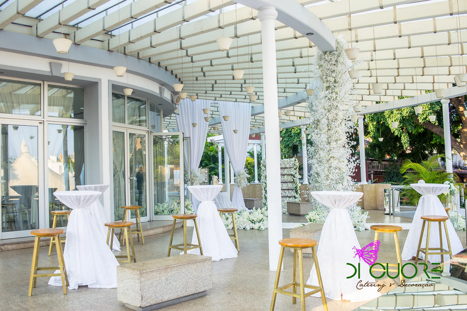 Terraço coberto com mesas altas de saia branca, bancos altos dourados e uma coluna de flores brancas