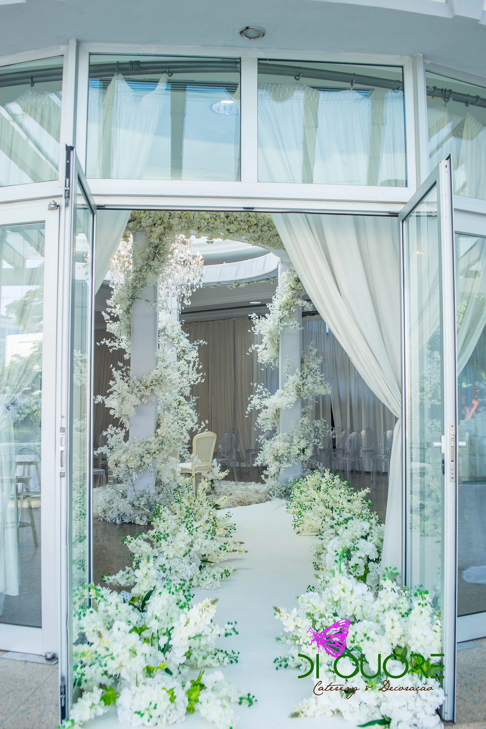 Portas de vidro abertas para o corredor branco de flores, com um cortinado e o pavilhão florido ao fundo