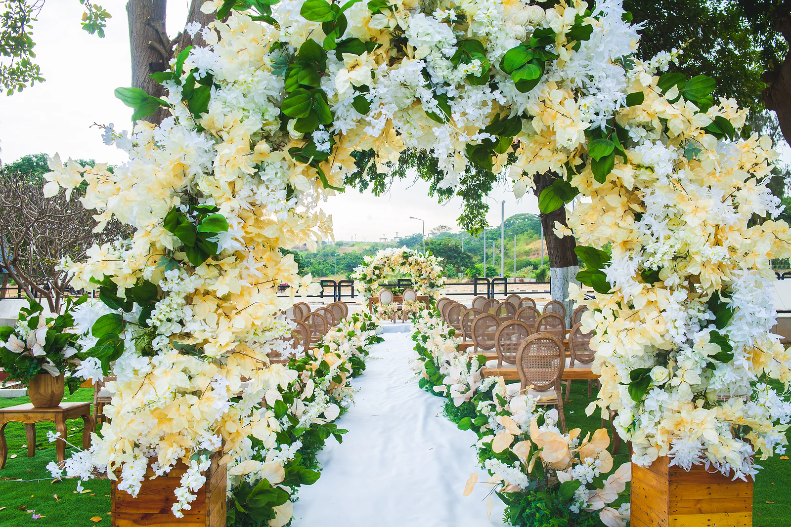 Passadeira branca a atravessar um arco de flores creme e folhagem, entre bancos de madeira e cadeiras de palhinha