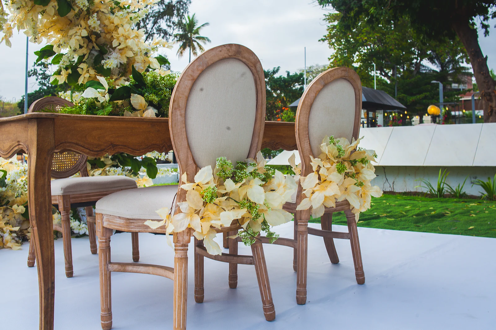 Duas cadeiras de madeira com costas de tecido, cada uma com um ramo de flores creme e folhagem, junto a uma mesa de madeira trabalhada