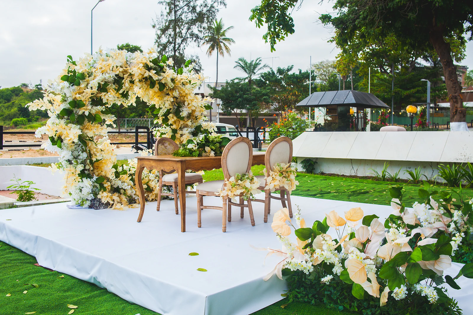 Estrado branco com um arco redondo de flores creme, uma mesa de madeira e duas cadeiras com ramos de flores, e flores creme em primeiro plano
