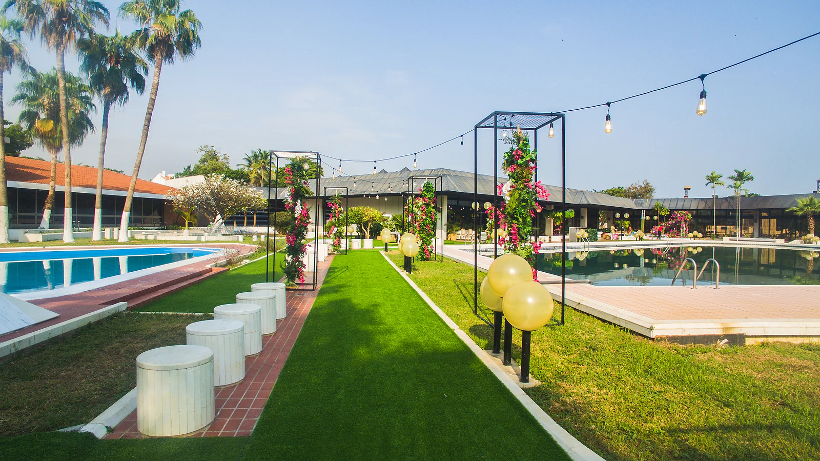 Relvado com estruturas de metal preto cobertas de flores, luzes em fio, bancos brancos redondos e a piscina ao fundo